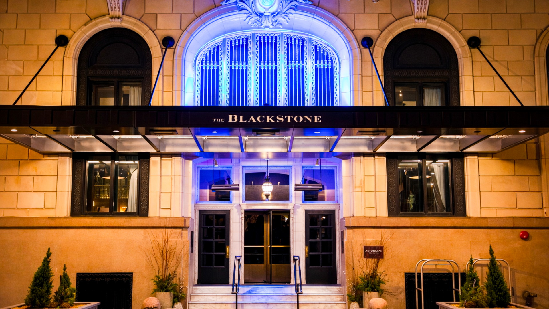 The exterior of The Blackstone building illuminated with blue lighting, featuring arched windows, decorative stonework, and a black awning over the entrance.