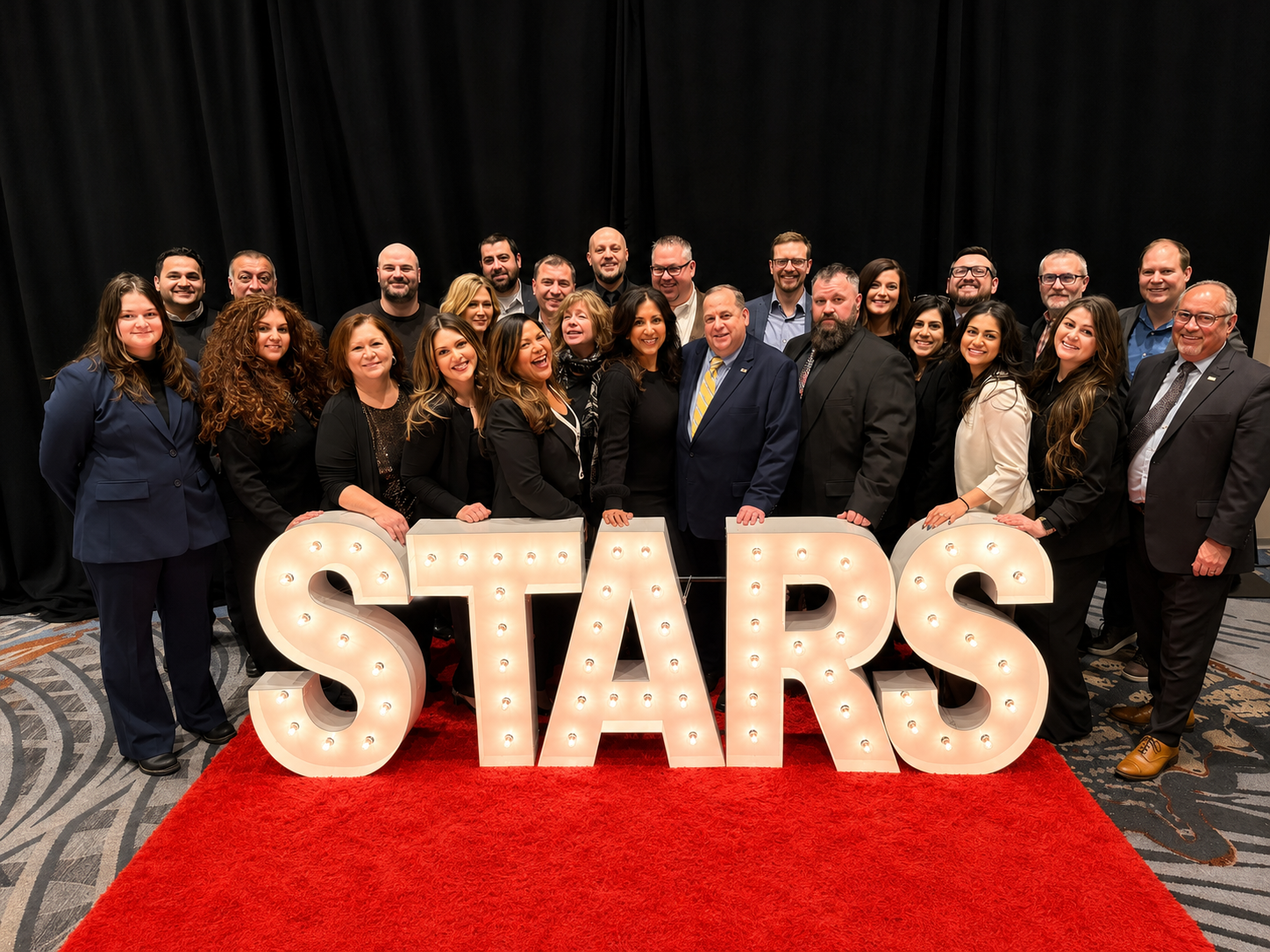 Group of diverse people at a corporate event posing behind illuminated 'STARS' sign on a red carpet with black curtains in the background.