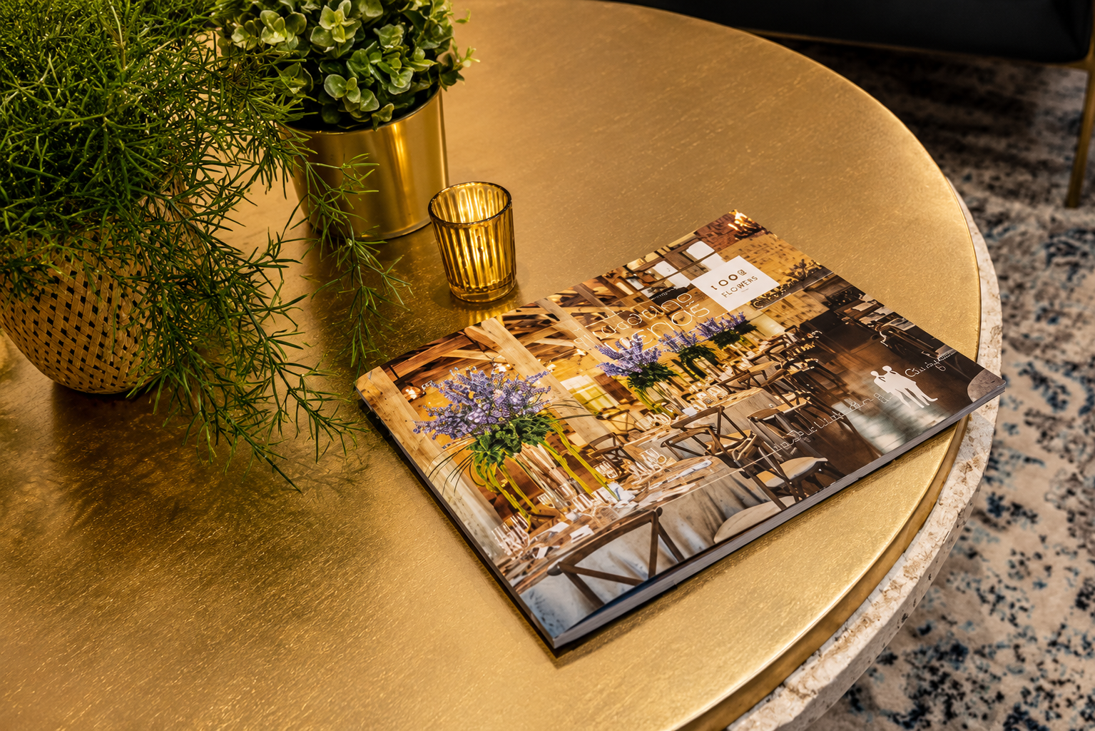 A round gold table with a potted plant, a small gold candle holder, and a catalog featuring a floral interior design on its cover.