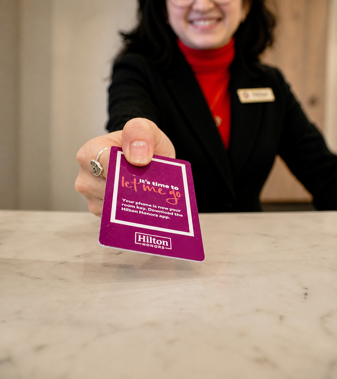 A woman in a black blazer and red turtleneck extending a purple Hilton Honors keycard towards the camera, with a smile on her face.
