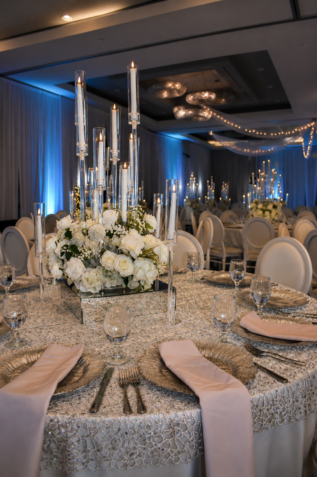 A decorated banquet table at an event, featuring a floral centerpiece with white roses and hydrangeas, surrounded by tall glass candle holders with lit candles, set with white napkins and elegant glassware, in a dimly lit room with blue accent lighting and additional tables with similar decor.