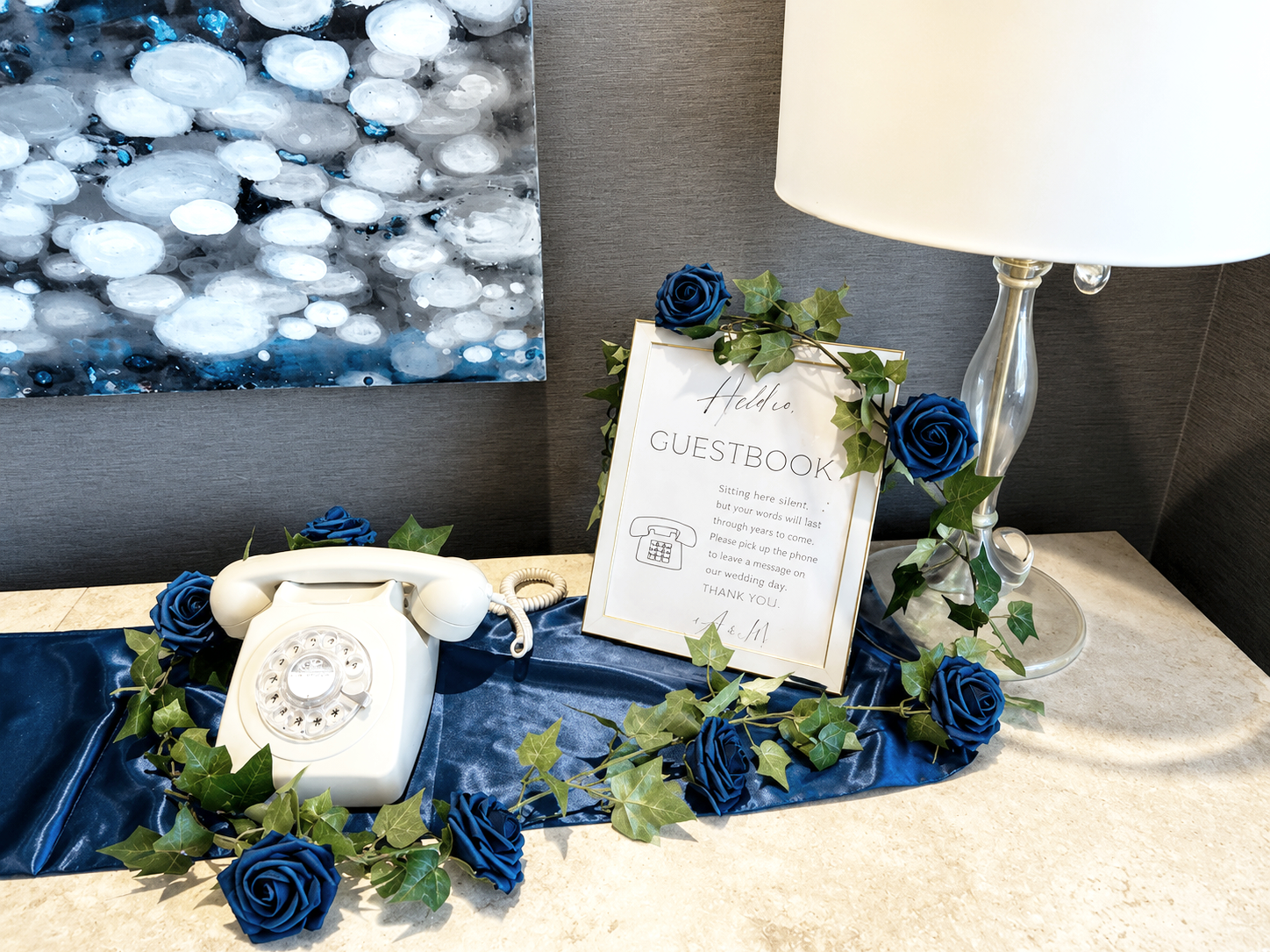 The image shows a white vintage rotary telephone with a spiral cord, placed on a navy blue satin cloth. Next to the telephone, there is a framed sign labeled 'Hello, GUESTBOOK,' with instructions for guests to leave messages on a phone. Blue roses and green ivy leaves decorate the area, and a large table lamp with a white shade and clear glass body is also visible. A backdrop includes a modern abstract painting with white, gray, and blue tones.