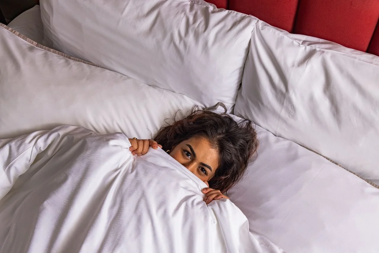 Woman with curly hair peeking from under white bedsheets in bed with multiple pillows