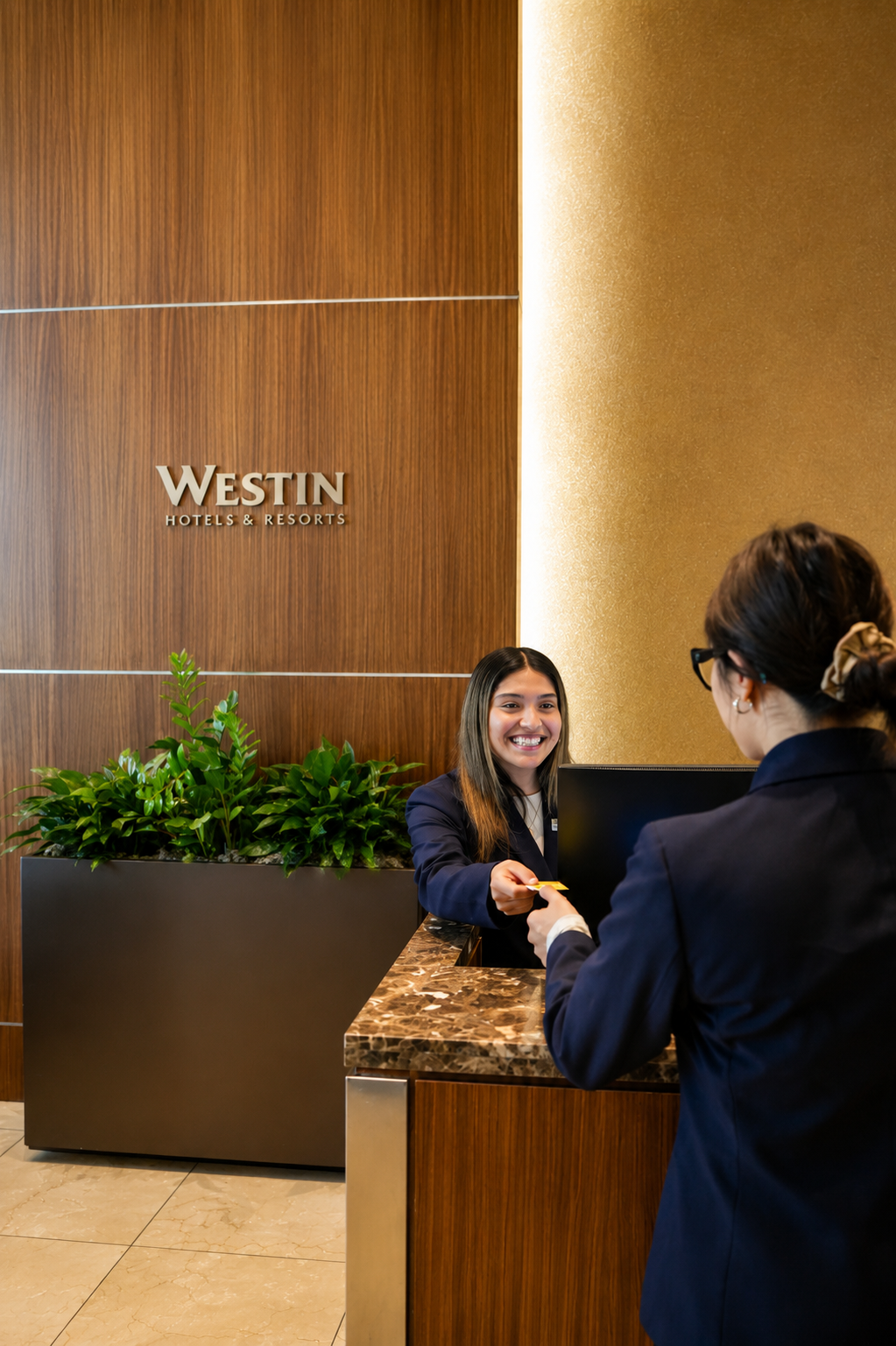 A hotel receptionist smiling at a guest at the Front Desk of the Westin Hotels & Resorts, with wooden wall paneling, a large green plant, and a marble countertop.