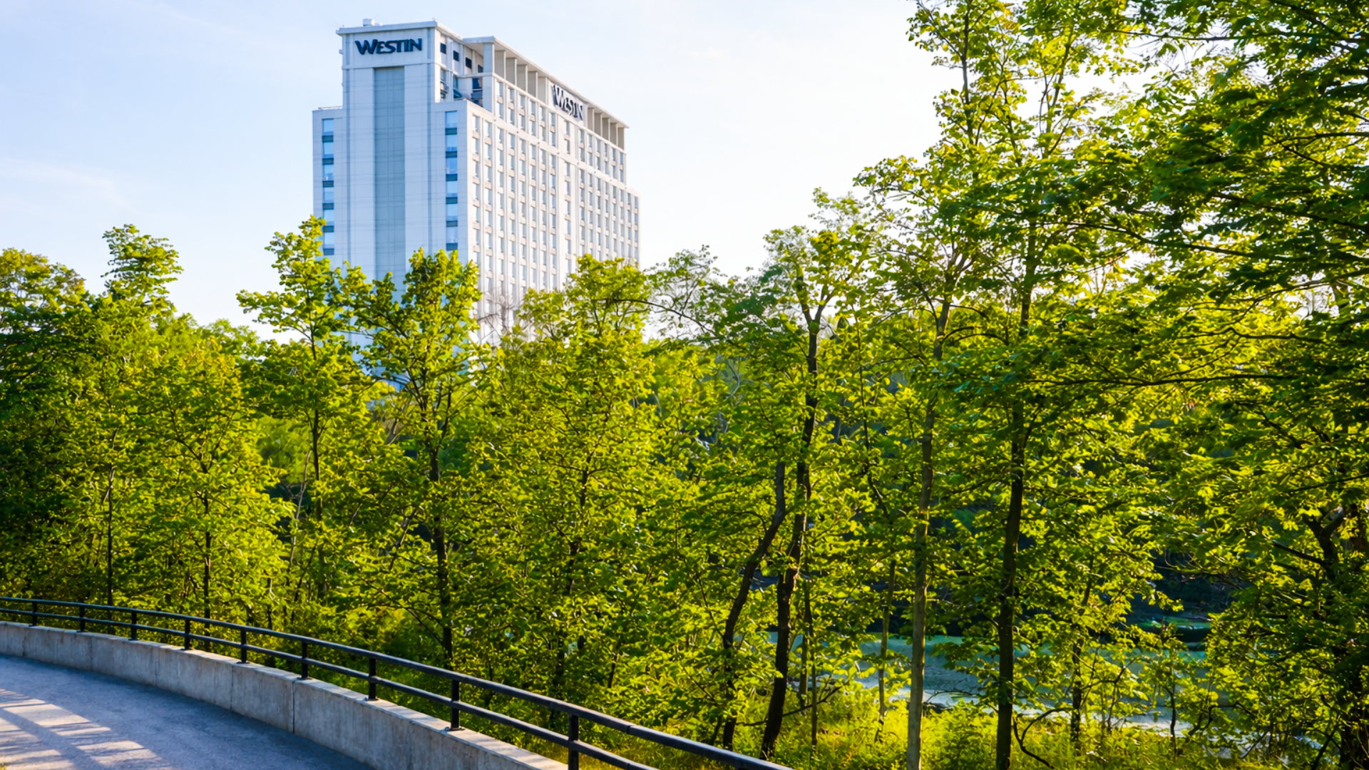 A tall modern building with the word 'Westin' on the top, seen behind lush green trees and a curved sidewalk with a metal railing in the foreground.