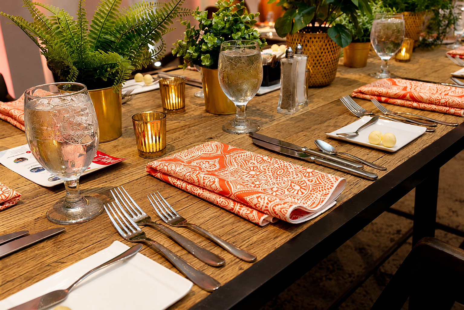 A styled dining table set with water glasses, gold and amber candles, green potted plants, orange and white patterned napkins, white square plates, silverware, small white cheese balls, and salt and pepper shakers, on a wooden table with warm ambient lighting.