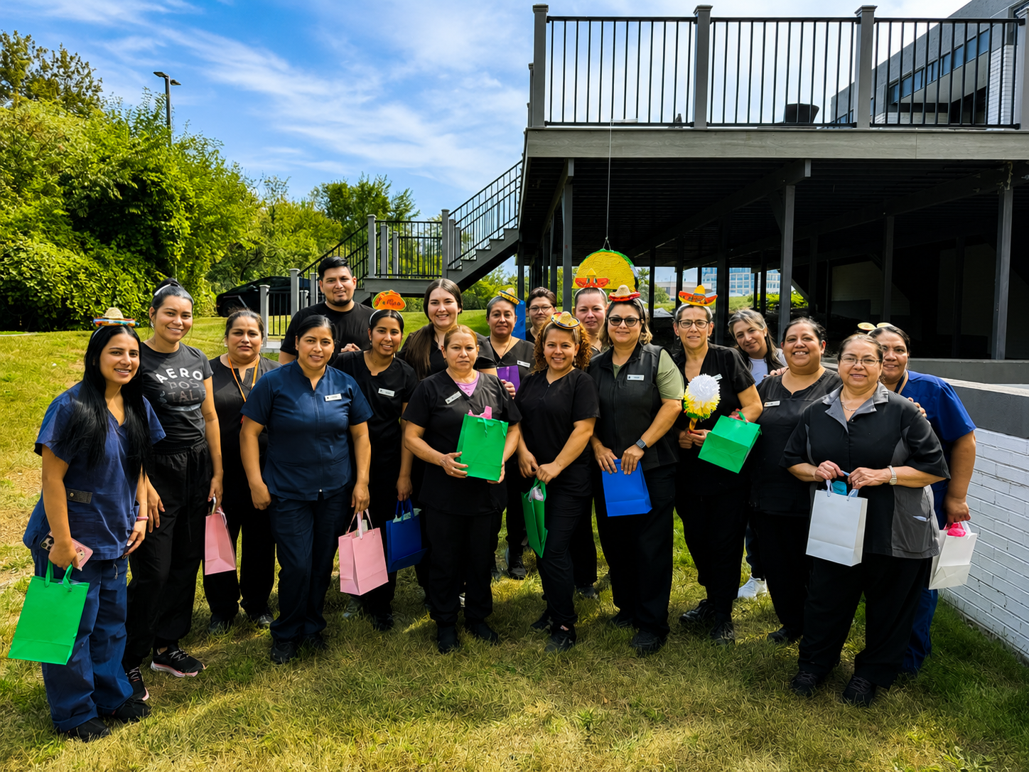 Group of healthcare workers standing outdoors, smiling, and holding gift bags and decorative items for a celebration. Some are wearing black scrubs, and a few have small festive hats.