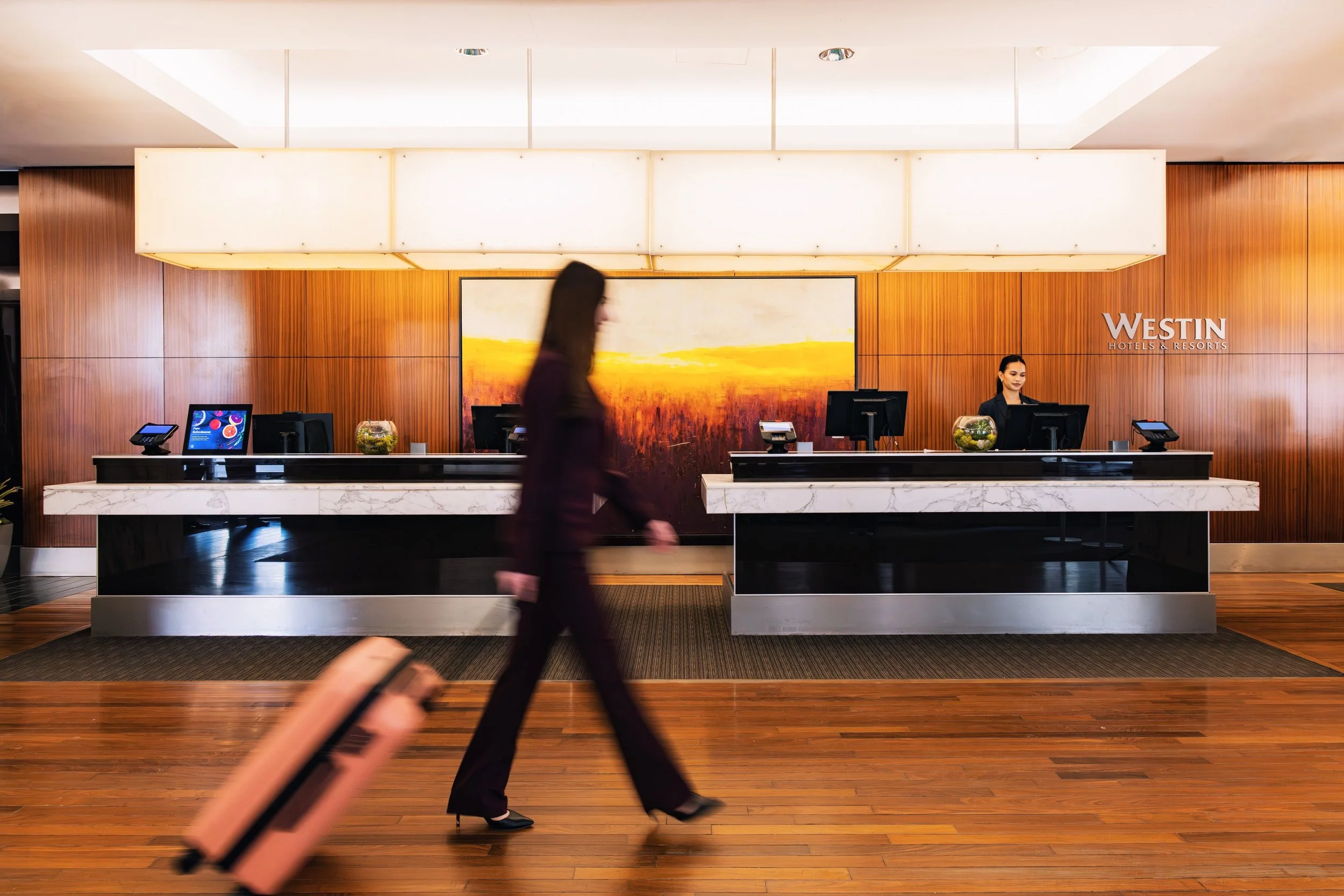 Lobby reception area at Westin Hotels & Resorts with a woman in business attire walking past the counter and a person holding a pink suitcase walking in the foreground.