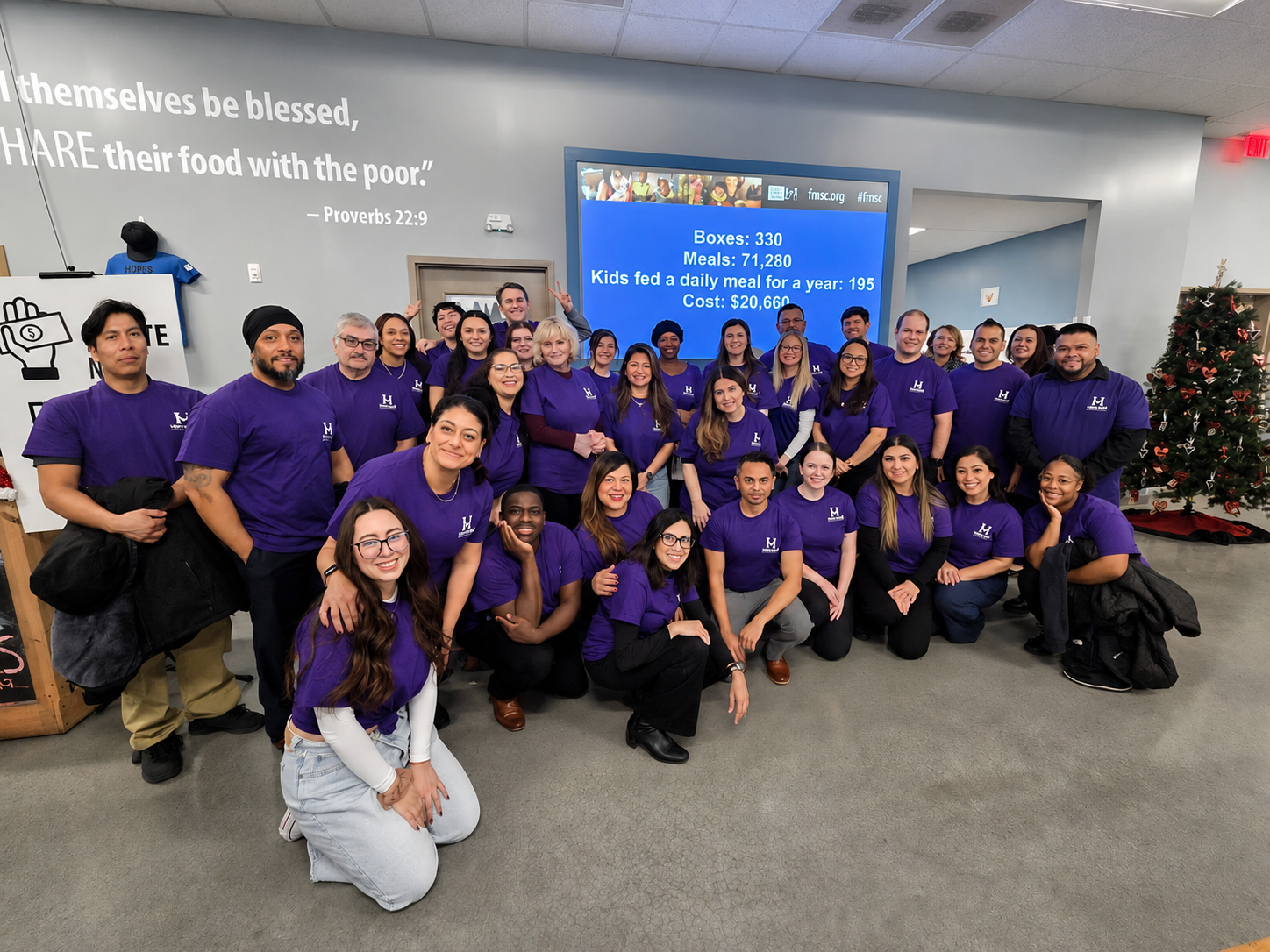 Group of volunteers wearing purple shirts posing for a photo inside a community center. There is a Christmas tree on the right and a large digital screen behind them displaying statistics about meals served. The wall has a Bible verse about sharing food with the poor.