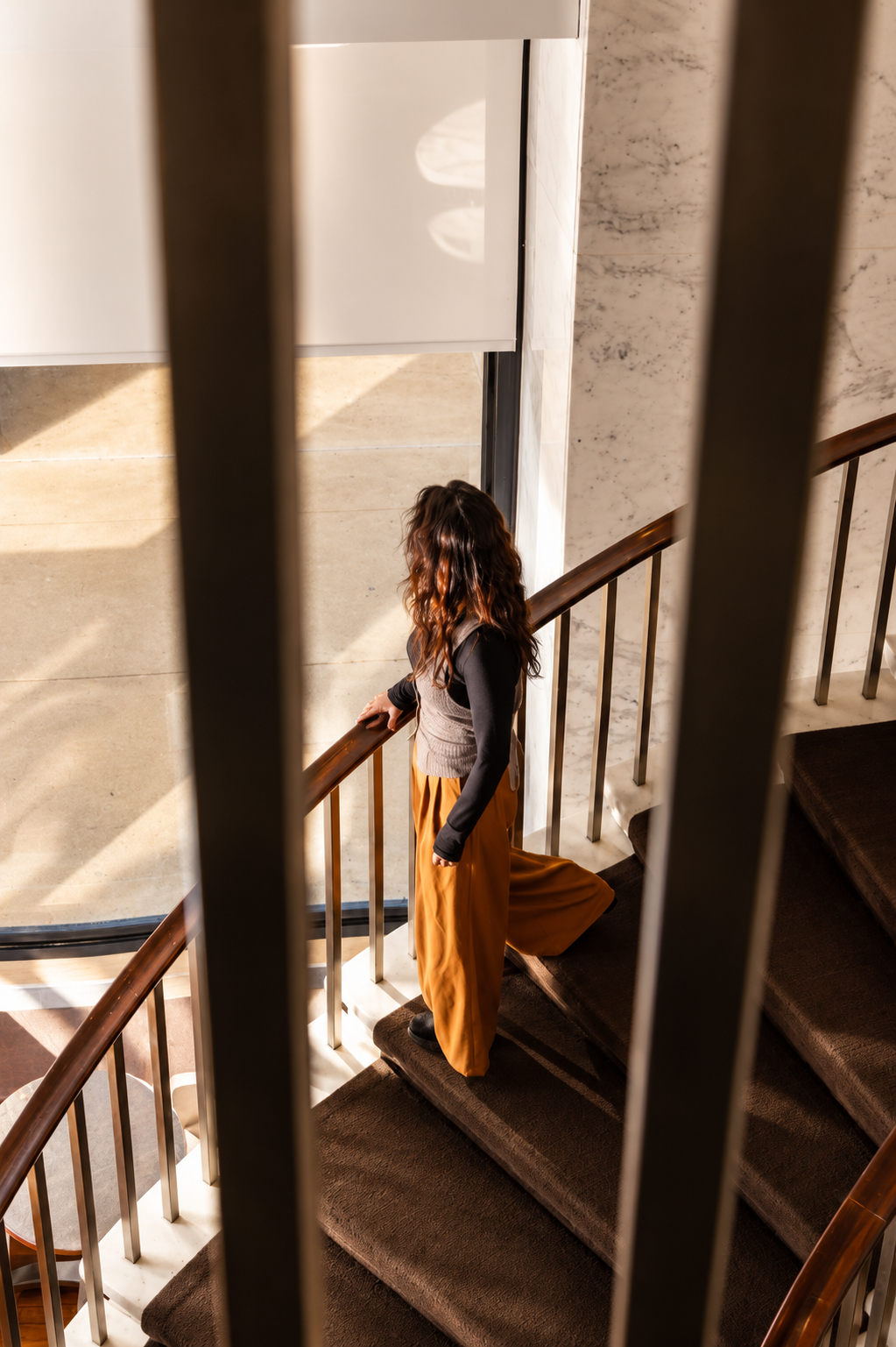 A woman with wavy hair wearing a black long sleeve top, a gray vest, and wide leg mustard-colored pants stands on a staircase, looking out of a glass door, with sunlight streaming into the space.