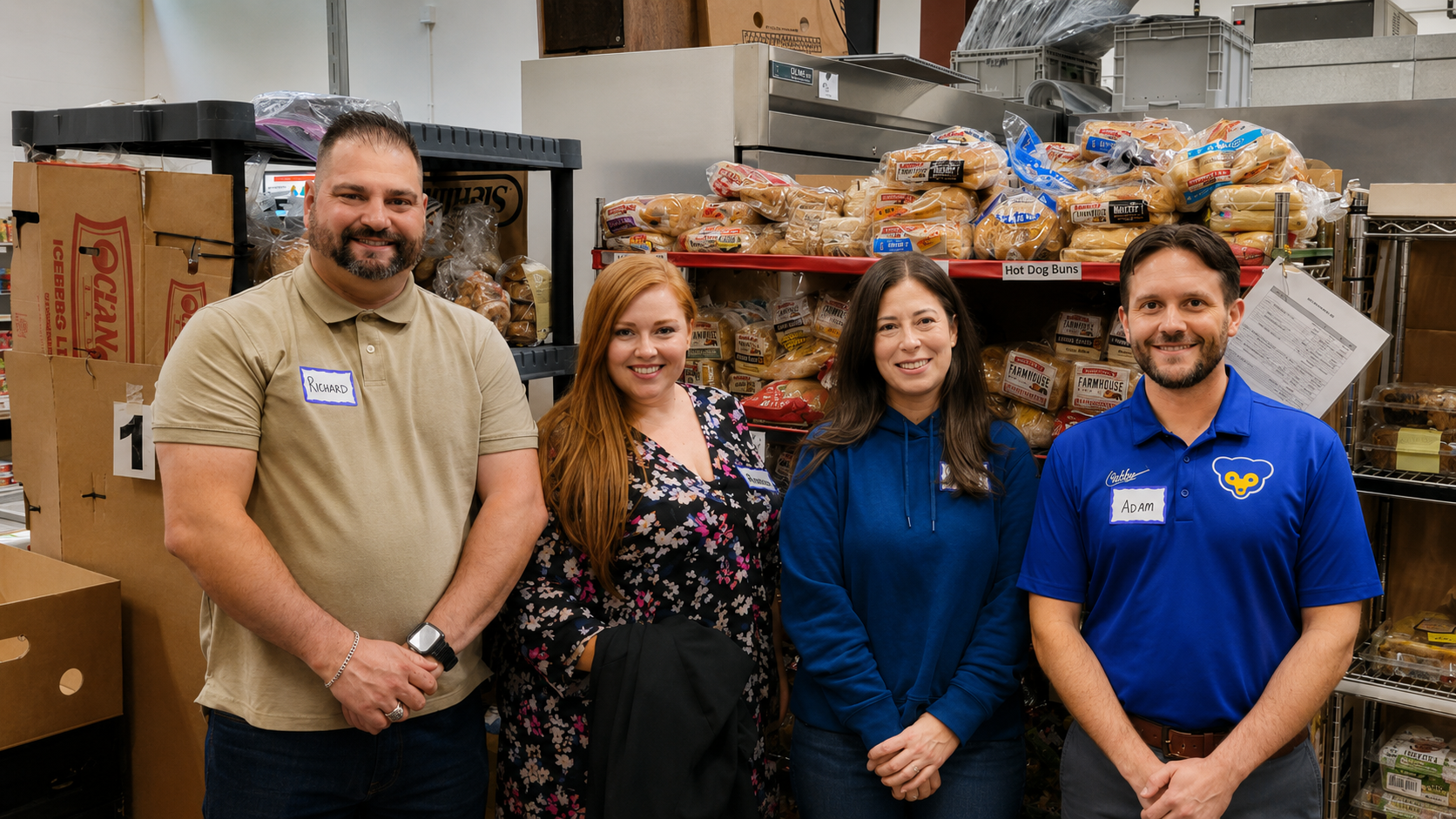 Four people standing in a storage room with shelves of bread and boxes behind them. Each person is smiling, with name tags on their shirts.