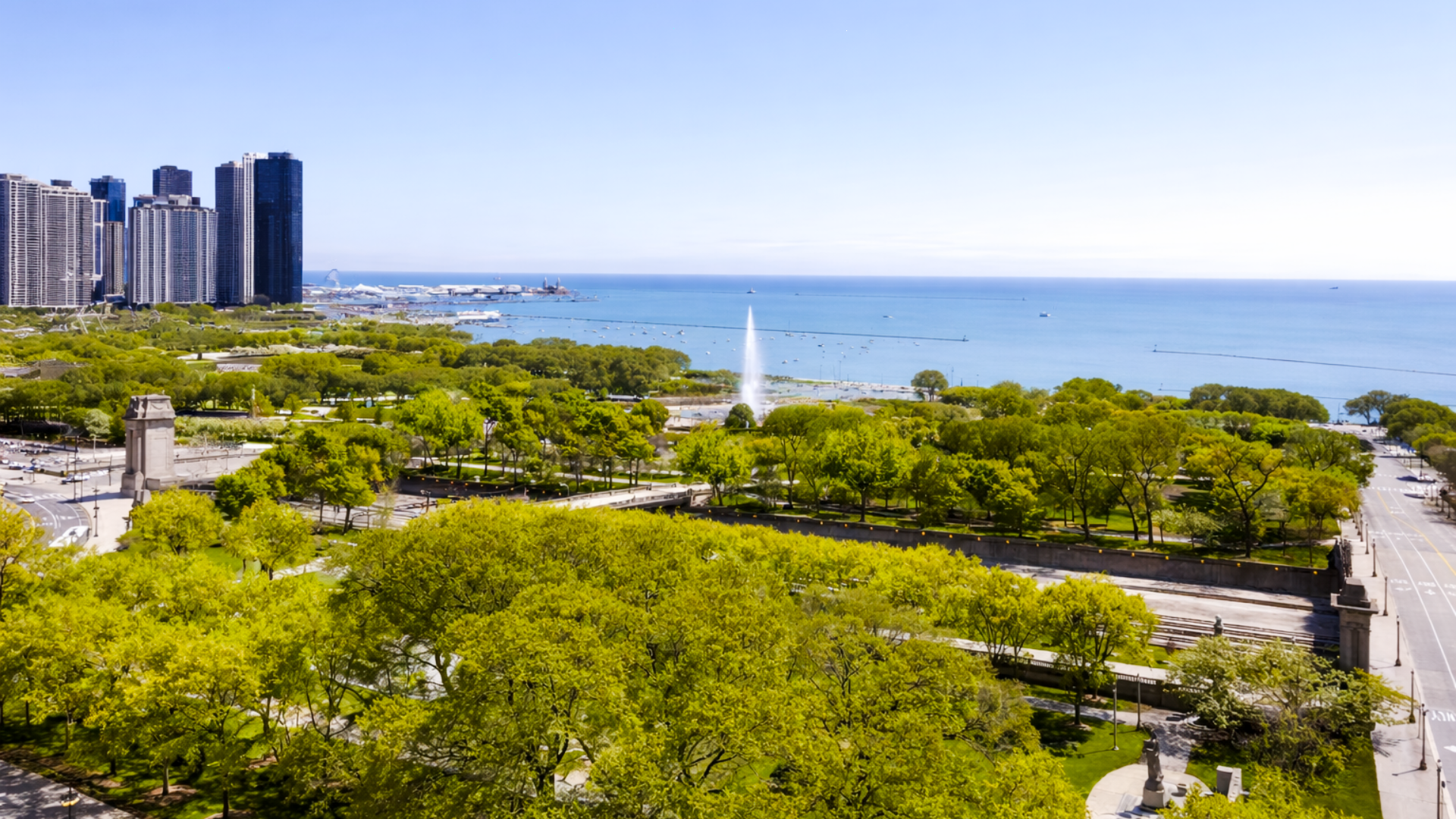 City skyline featuring tall modern buildings, a large park with lush green trees, a fountain in the middle, and the ocean in the background.
