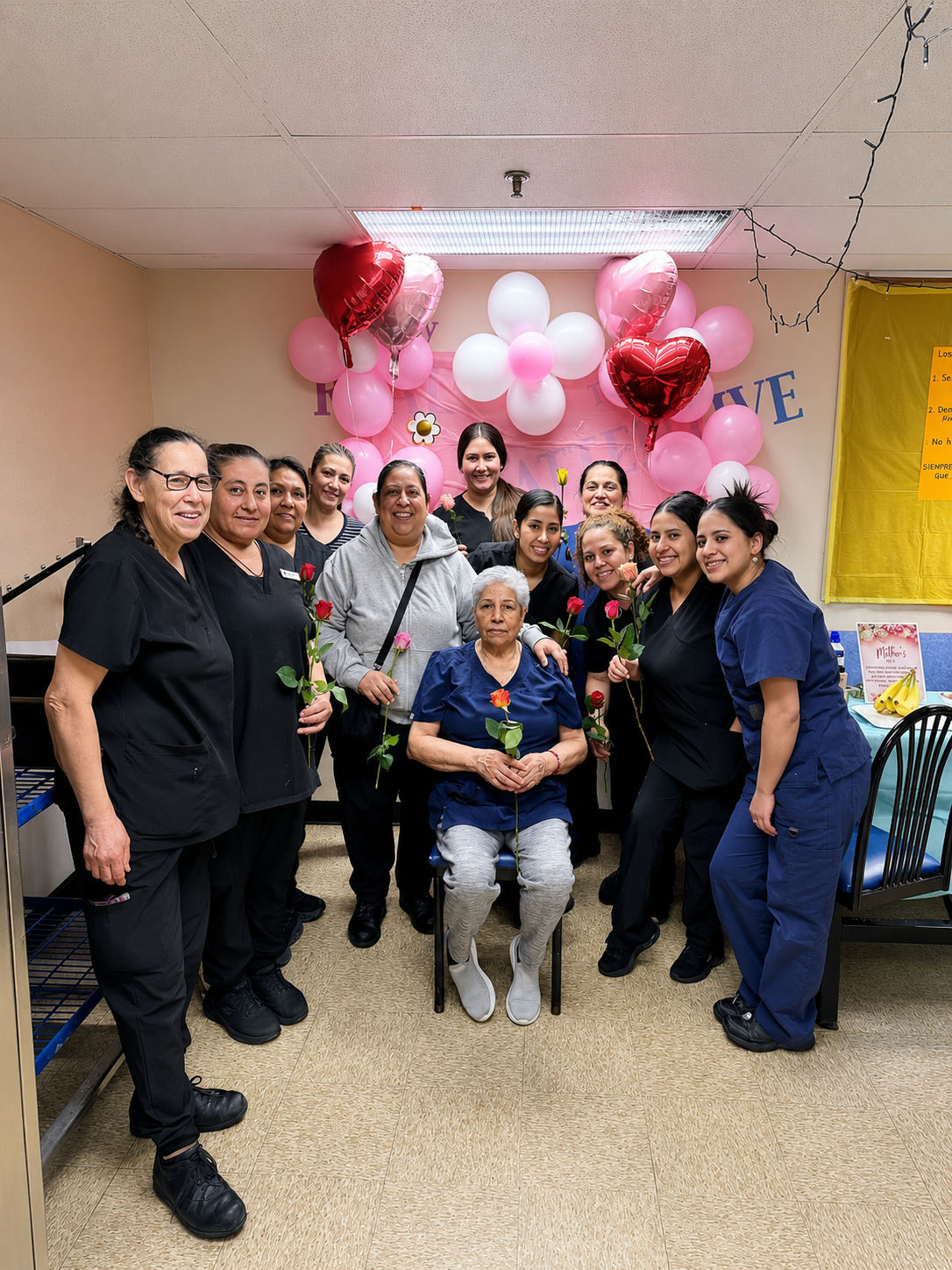 Group of women and an elderly woman celebrating with flowers and balloons in a decorated room.