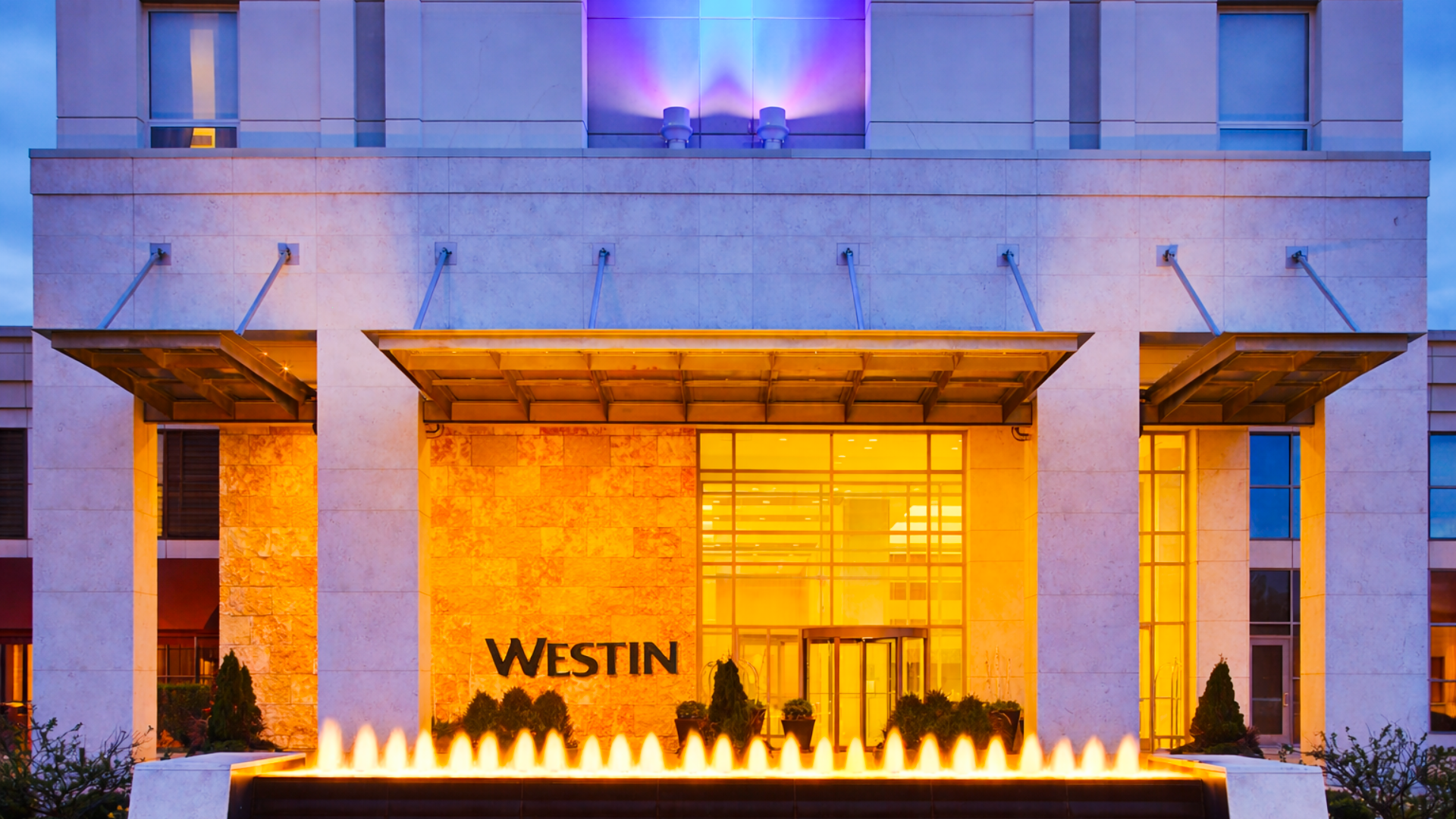 The exterior of a modern hotel building at sunset with a fountain in front, illuminated with colorful purple, orange, and blue lights, and the word 'Westin' visible near the entrance.