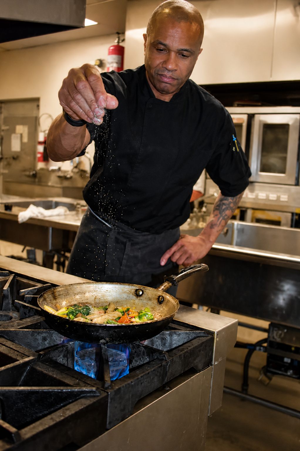 A chef sprinkling seasoning into a skillet with vegetables on a blue flame stove in a professional kitchen.