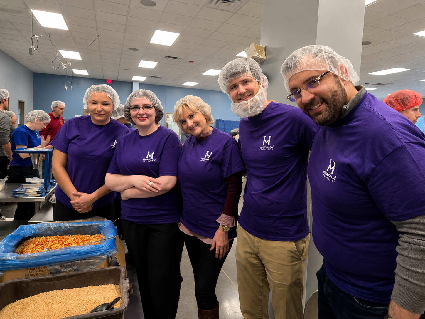 Group of five volunteers in purple shirts and hairnets standing together in a food packing event, with food trays and supplies on the table in front of them.