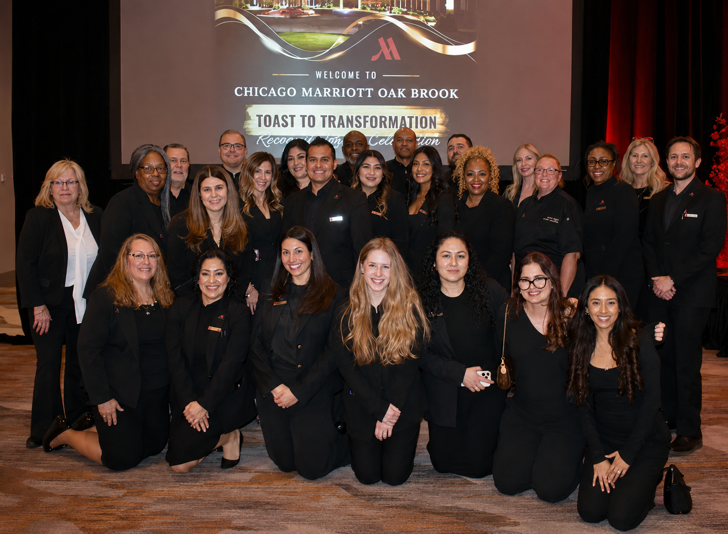 Group of people in business attire posing for a photo at a hotel event with a screen displaying ‘Welcome to Chicago Marriott Oak Brook’ in the background.