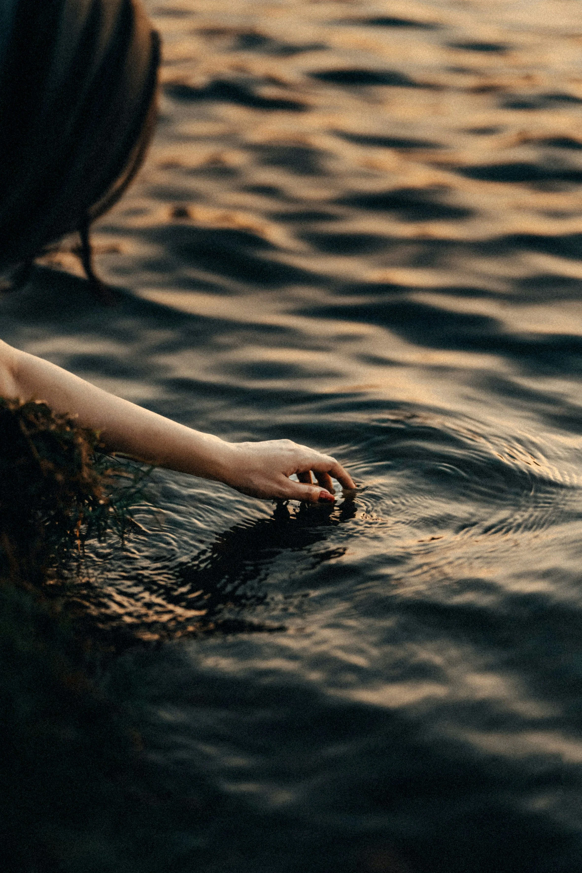 A person's arm extends from a dark outfit reaching into the water of a lake or river at sunset, with gentle ripples and warm tones reflecting on the surface.
