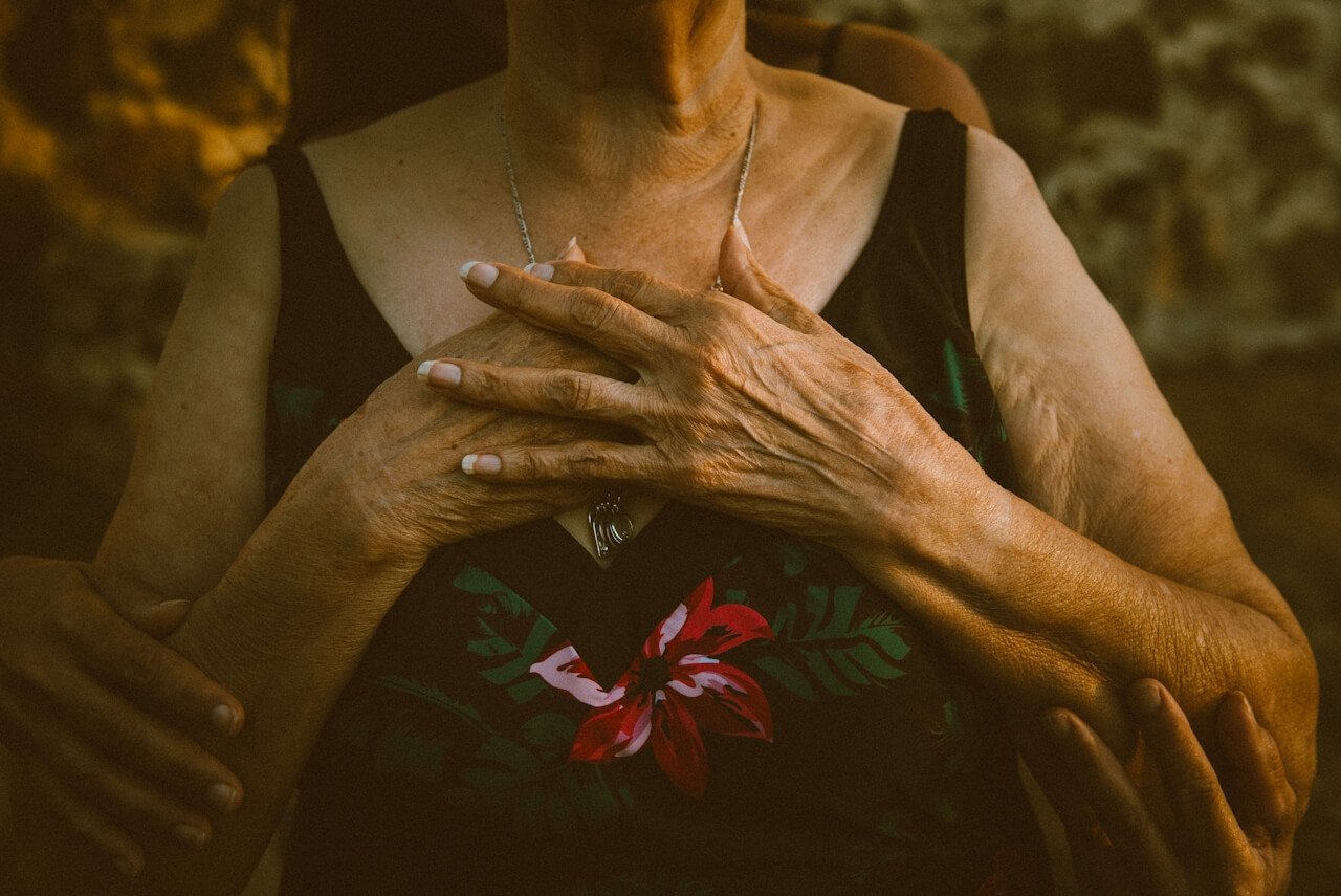 Close-up of an elderly woman with tanned skin and white nails, wearing a black top with a red and white floral pattern, holding her hands over her chest, with multiple hands of different sizes gently touching her.