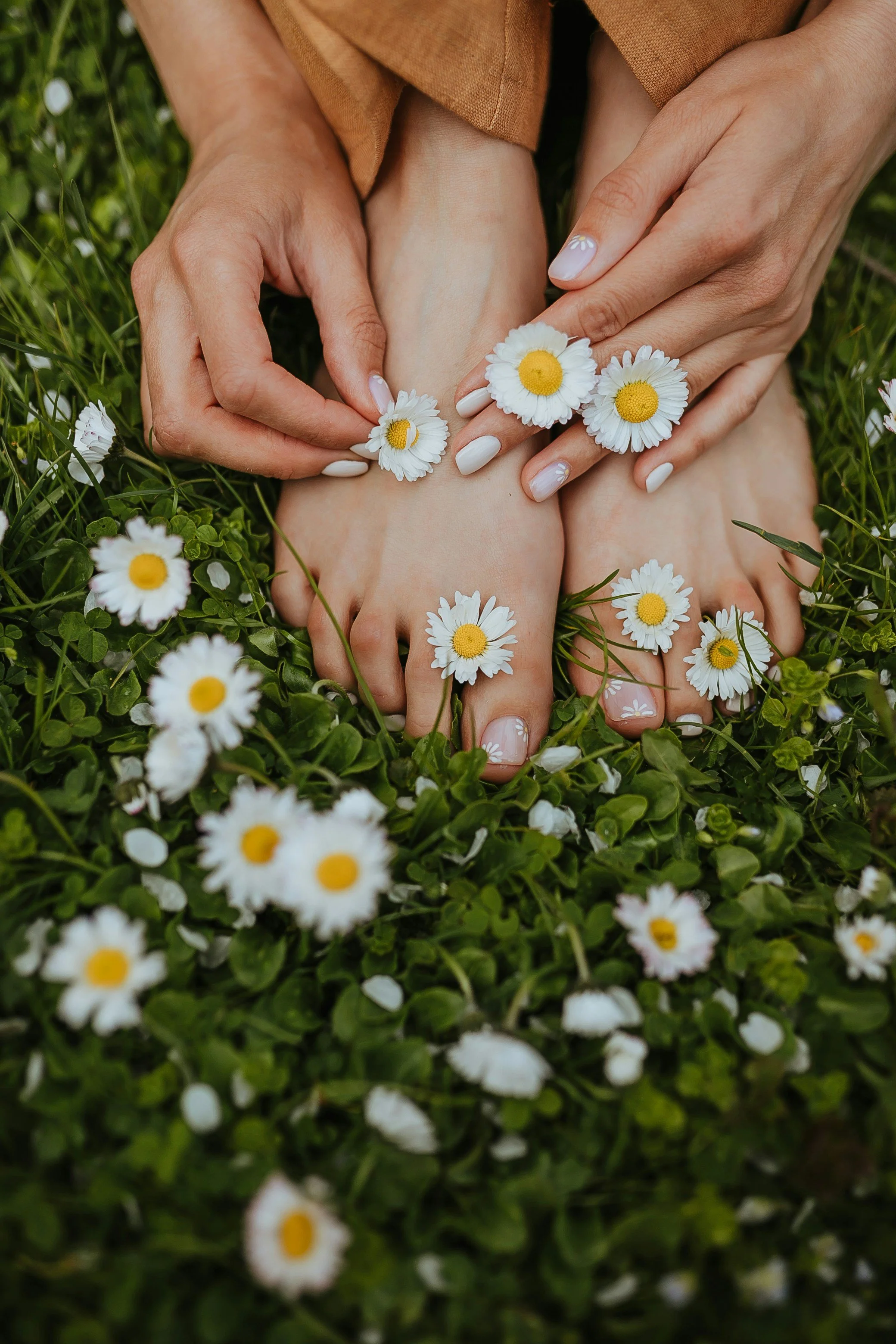Close-up of a person's hands holding two feet, decorated with daisies and surrounded by more daisies in grass.