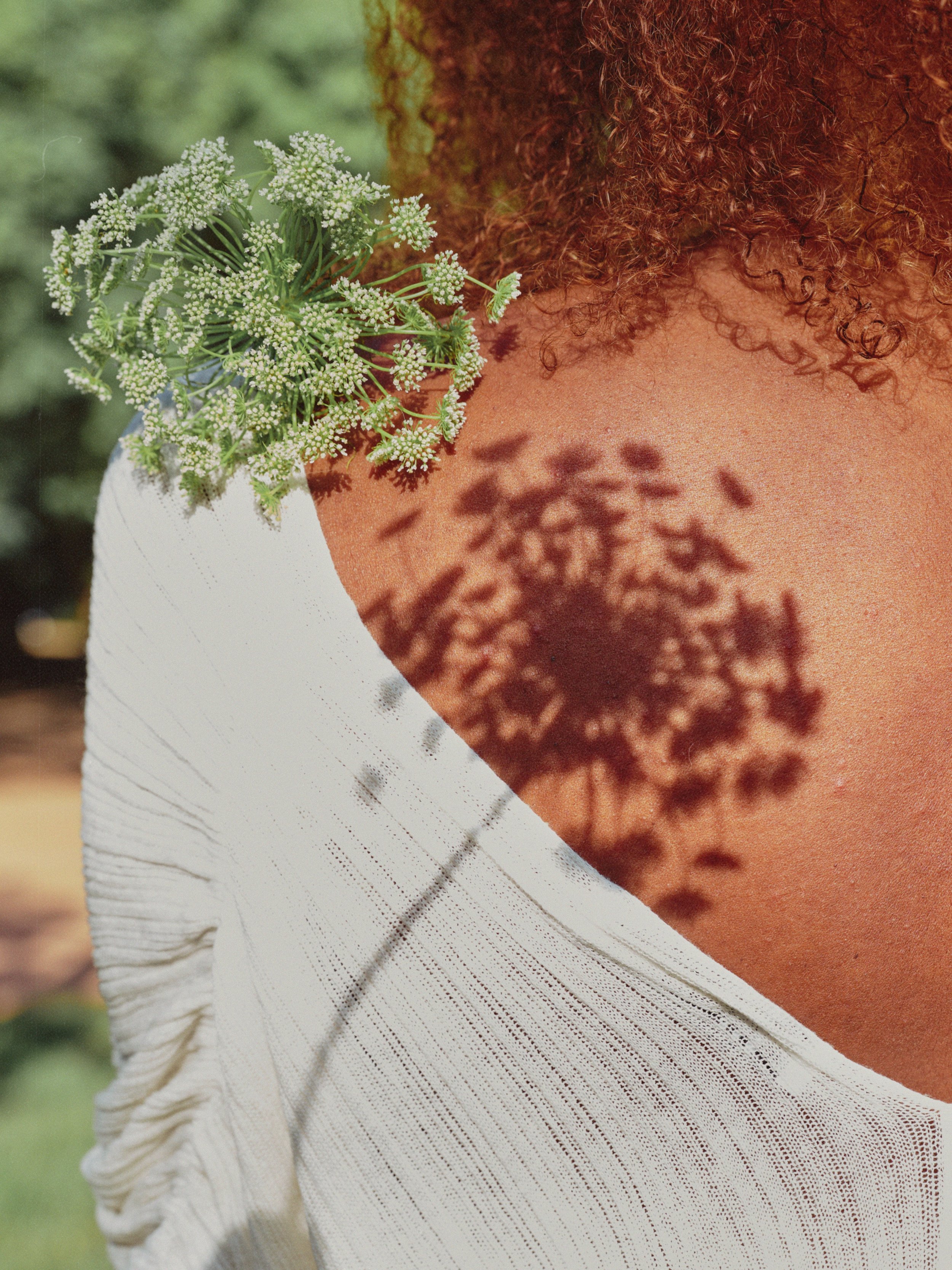 Close-up of a person with curly red hair and brown skin, holding a bunch of white flowers with shadow pattern on their shoulder, wearing a white textured garment.