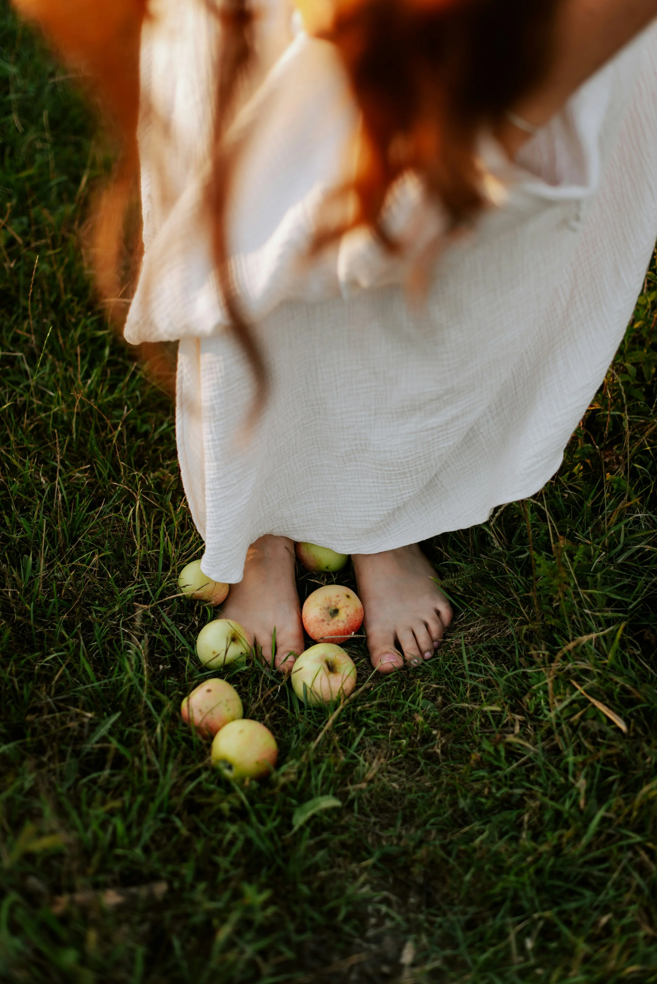 A person standing on grass wearing a long white dress with apples scattered around their bare feet.