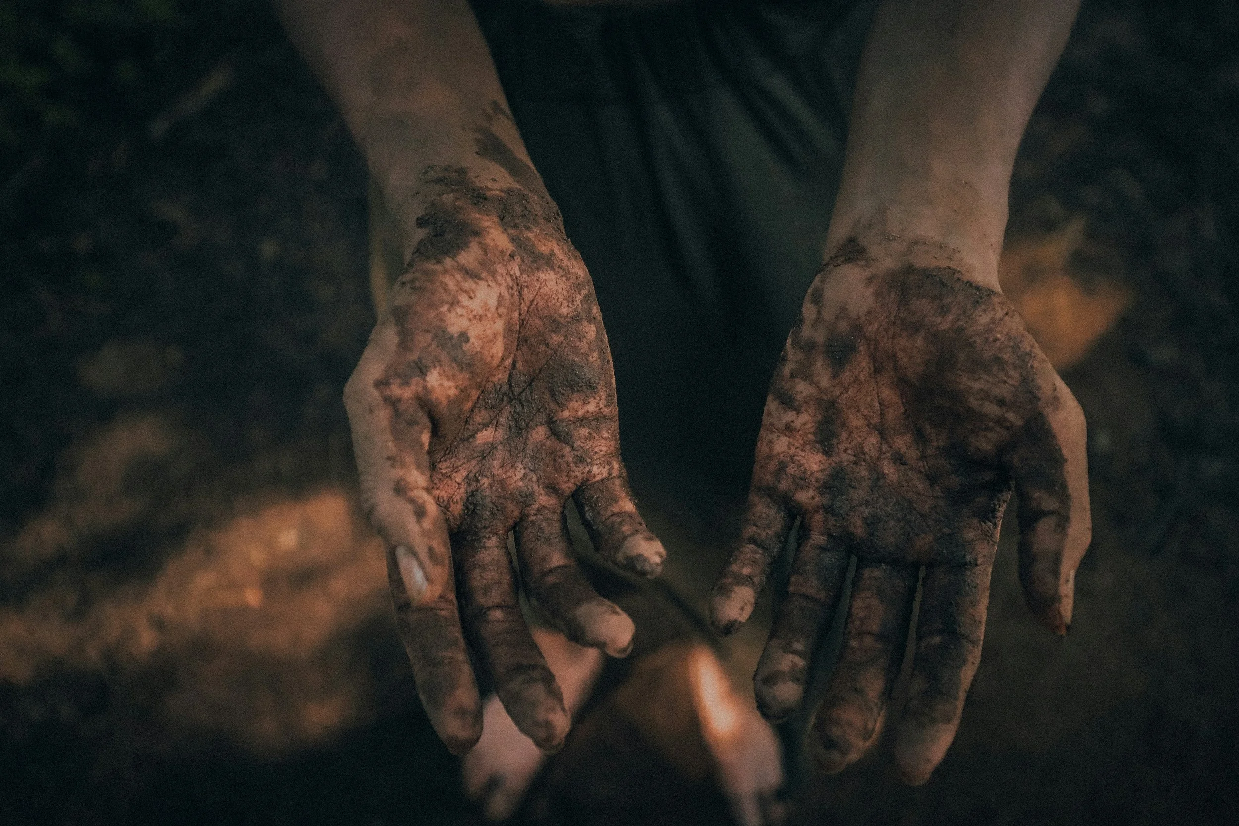 Dirty hands with soil, holding a tool with a glowing ember in a dark setting.
