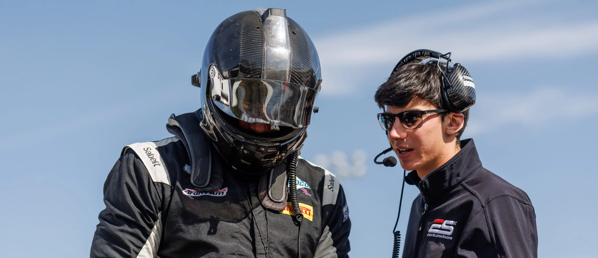 Two men, one in a racing suit and helmet, and the other in a black racing jacket with a headset and microphone, talking outdoors on a sunny day against a blue sky.