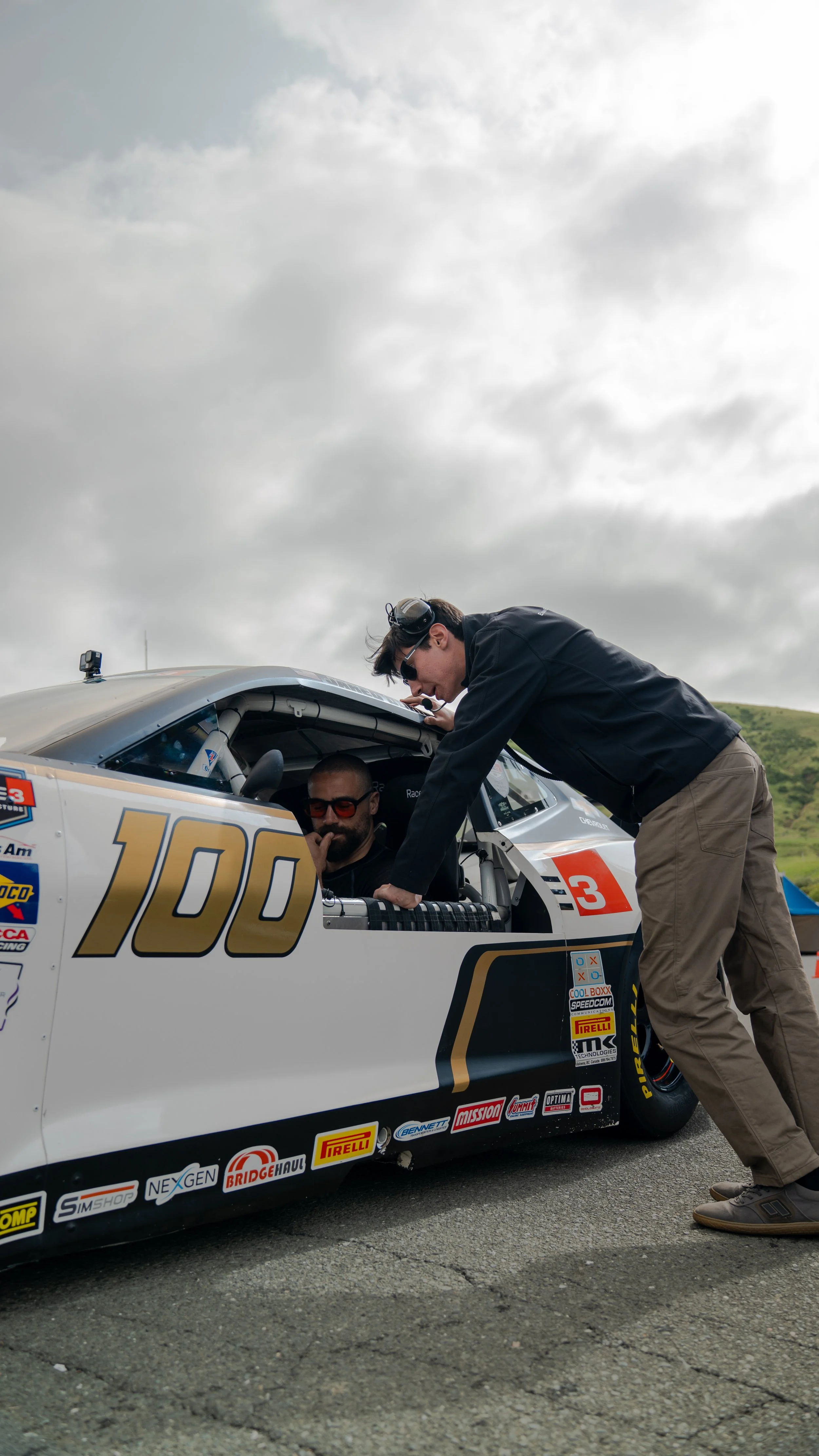 A man in black jacket and khaki pants leaning into a race car with a driver inside, who is wearing sunglasses, at a race track on a cloudy day.