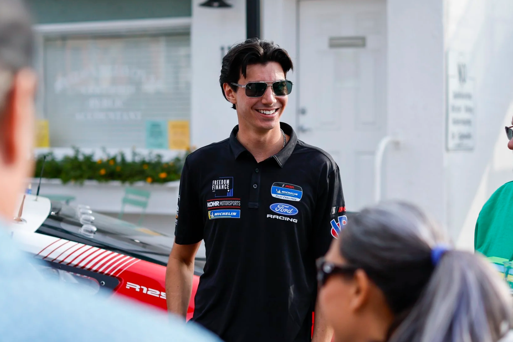 A young man smiling wearing sunglasses and a black racing shirt with sponsor logos, standing outdoors near a race car, in a social setting