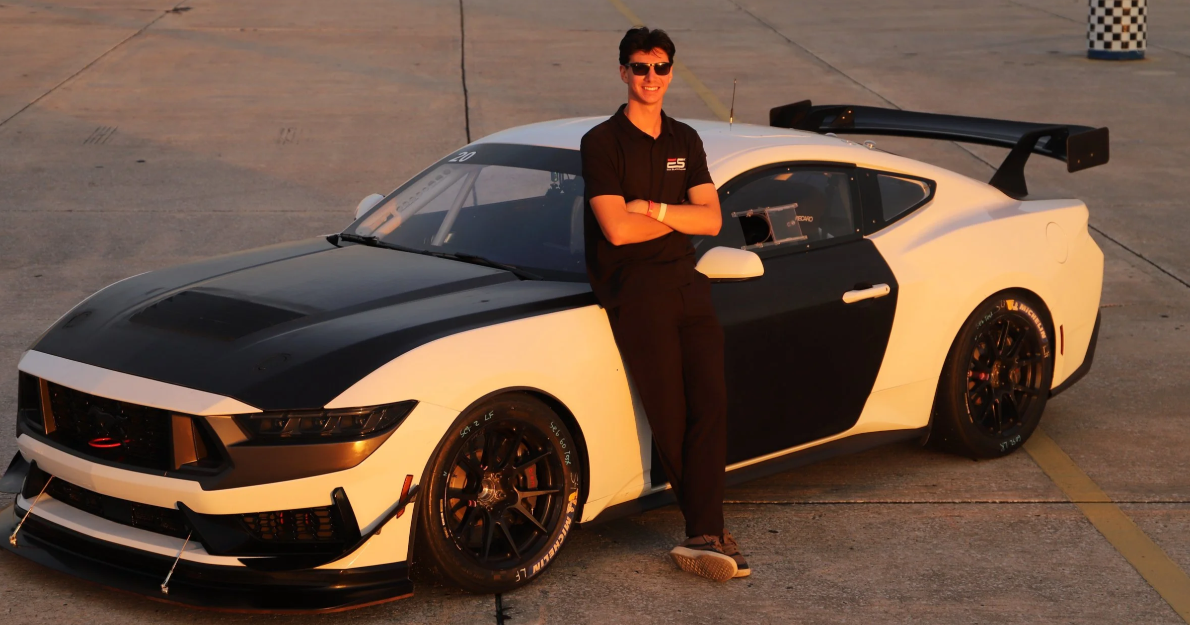 A young man in black clothing and sunglasses leaning against a white and black race car on a concrete surface, with a checkered parking lot cone in the background, during sunset.