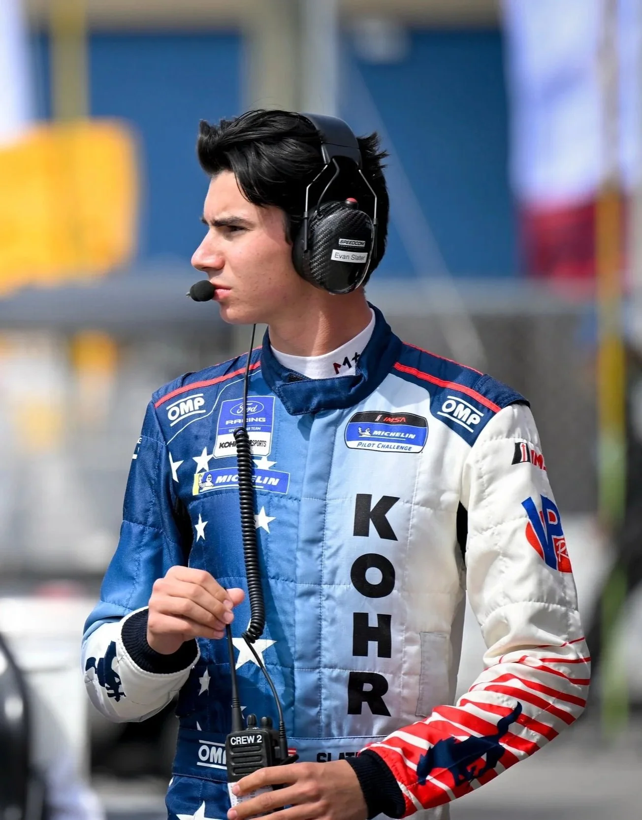 Race car driver in racing suit and headset, standing outdoors.