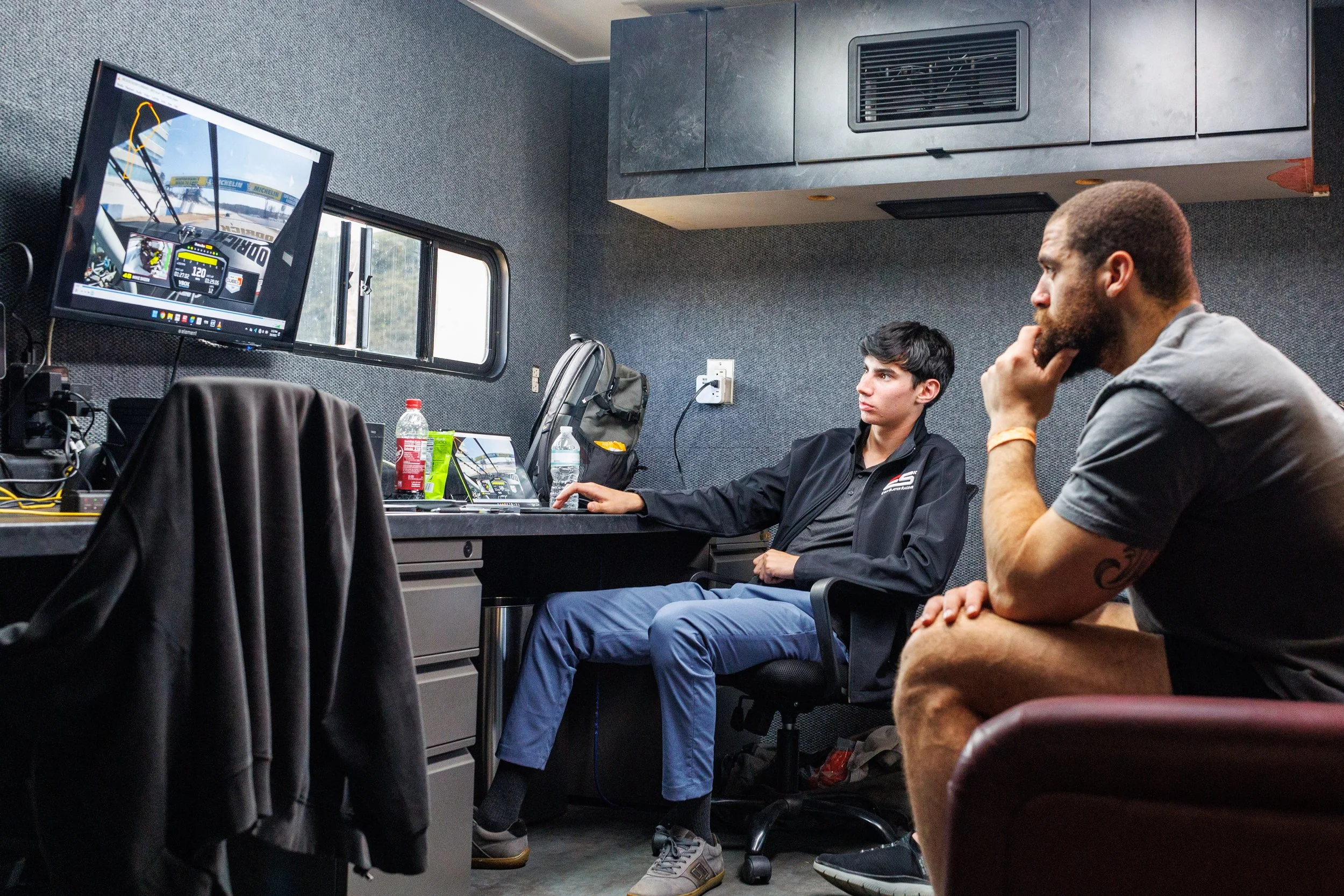 Two men in a room with a racing simulator on a computer monitor, focusing on a racing game or data analysis, with drinks and gear on the desk