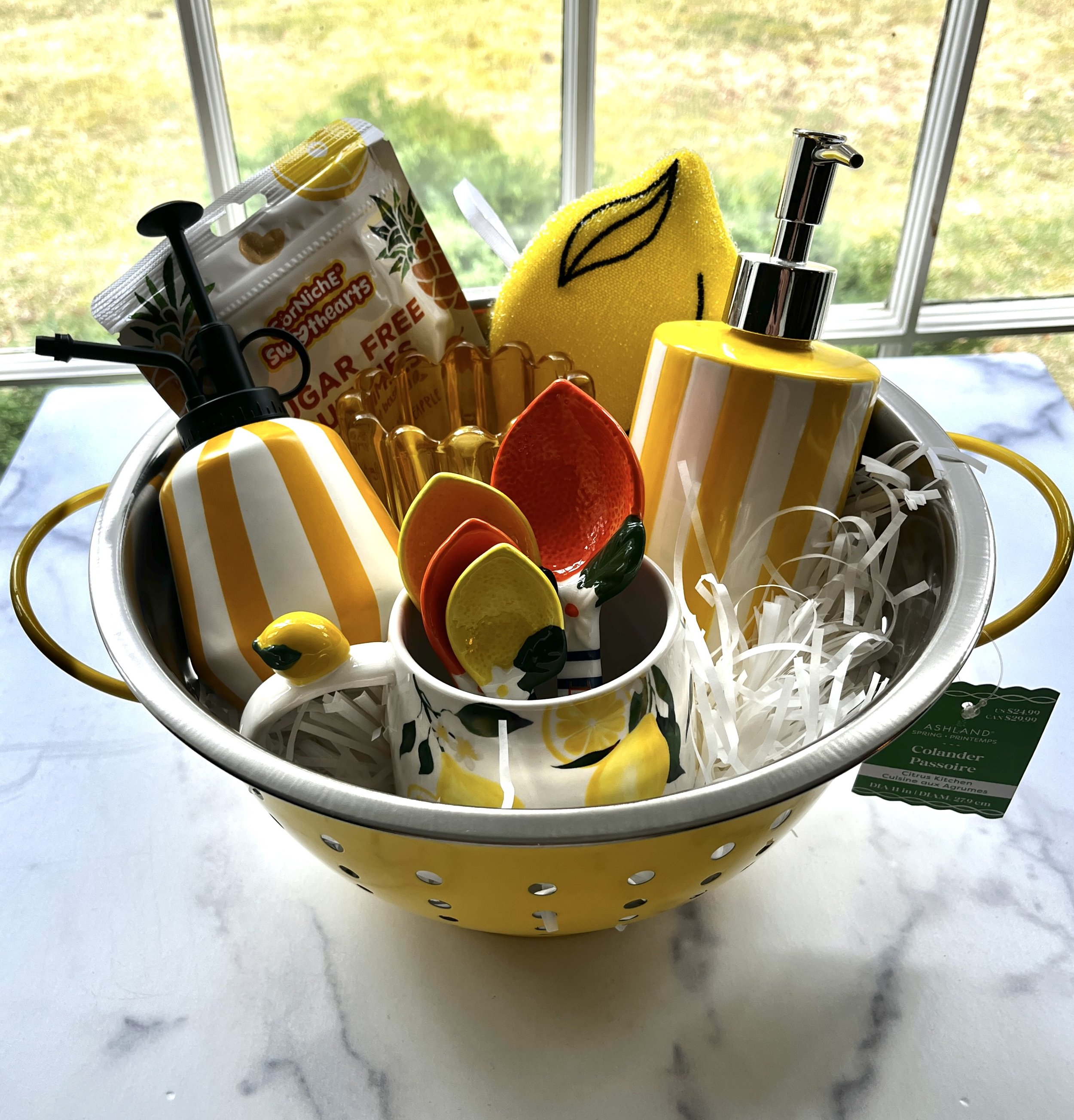 A lemon themed basket but instead of the items being in a basket they are in a yellow collanger with handles. Items inside the collander include a lemon scrubber, lemon teaspoons, a lemon mug, a yellow glass candle holder, a yellow soap dispenser etc