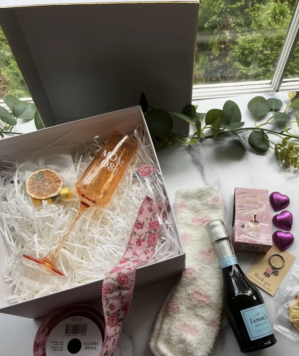 Image is of an opened white gift box with white paper shredded filler paper, with dried orange slices, a champagne glass that states "MOM-osa", a rose drink stirrer, floral ribbon, cozy women's socks, chocolates. All the items that come in the gift.
