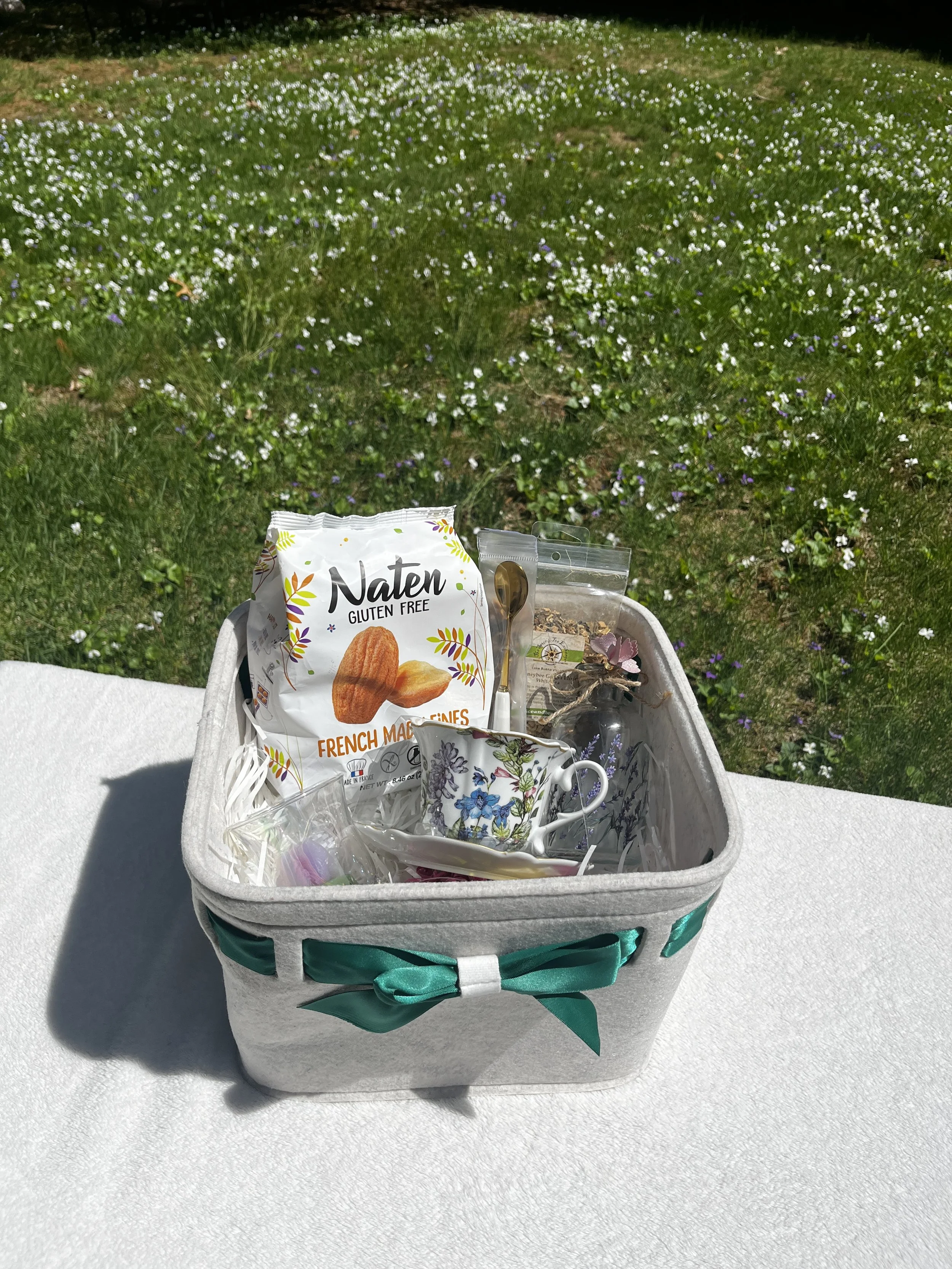 A tea time basket with a felt basket and dark green ribbon, Gluten free madeleine cookies, a tea cup and saucer, a votive tulip shaped candle, lavender salts in a reusable glass jar with lavender print.