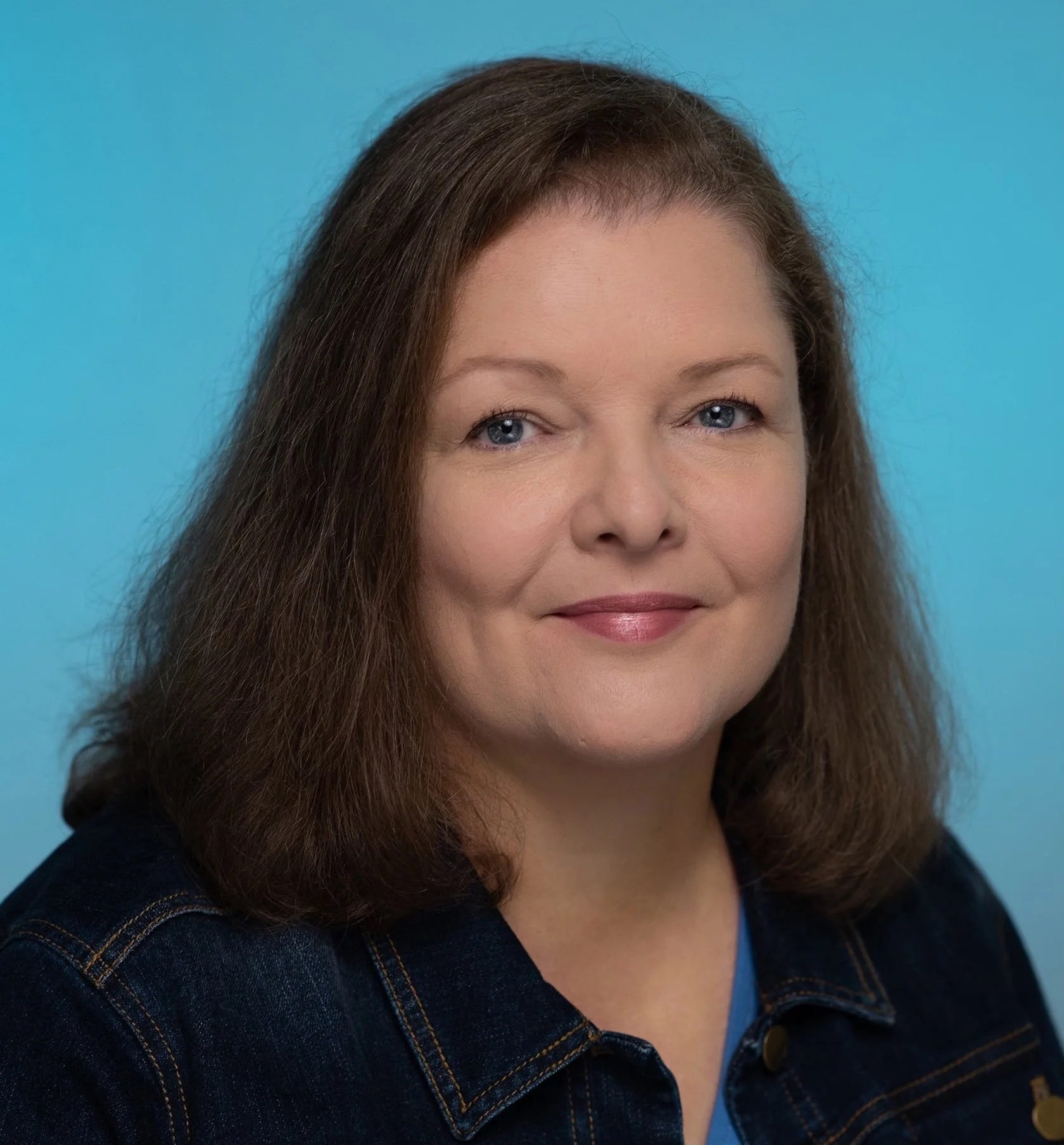 A color headshot showing a white woman with blue eyes and brown shoulder-length hair wearing a jean jacket and blue T-=shirt against a bright blue background.