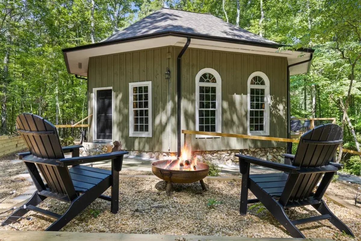 Cozy backyard fire pit area at a CoHost Works vacation rental with Adirondack chairs, a green cottage, gravel patio, and wooded surroundings.