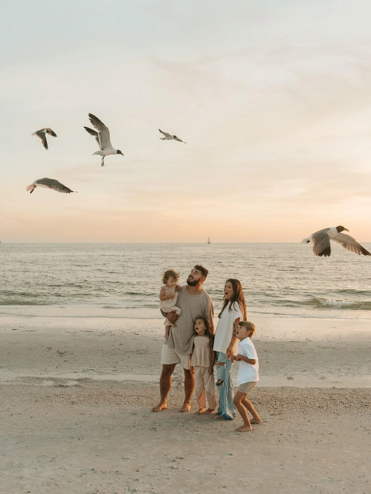 Family on a beach at sunset with seagulls flying overhead and a sailboat in the distance.