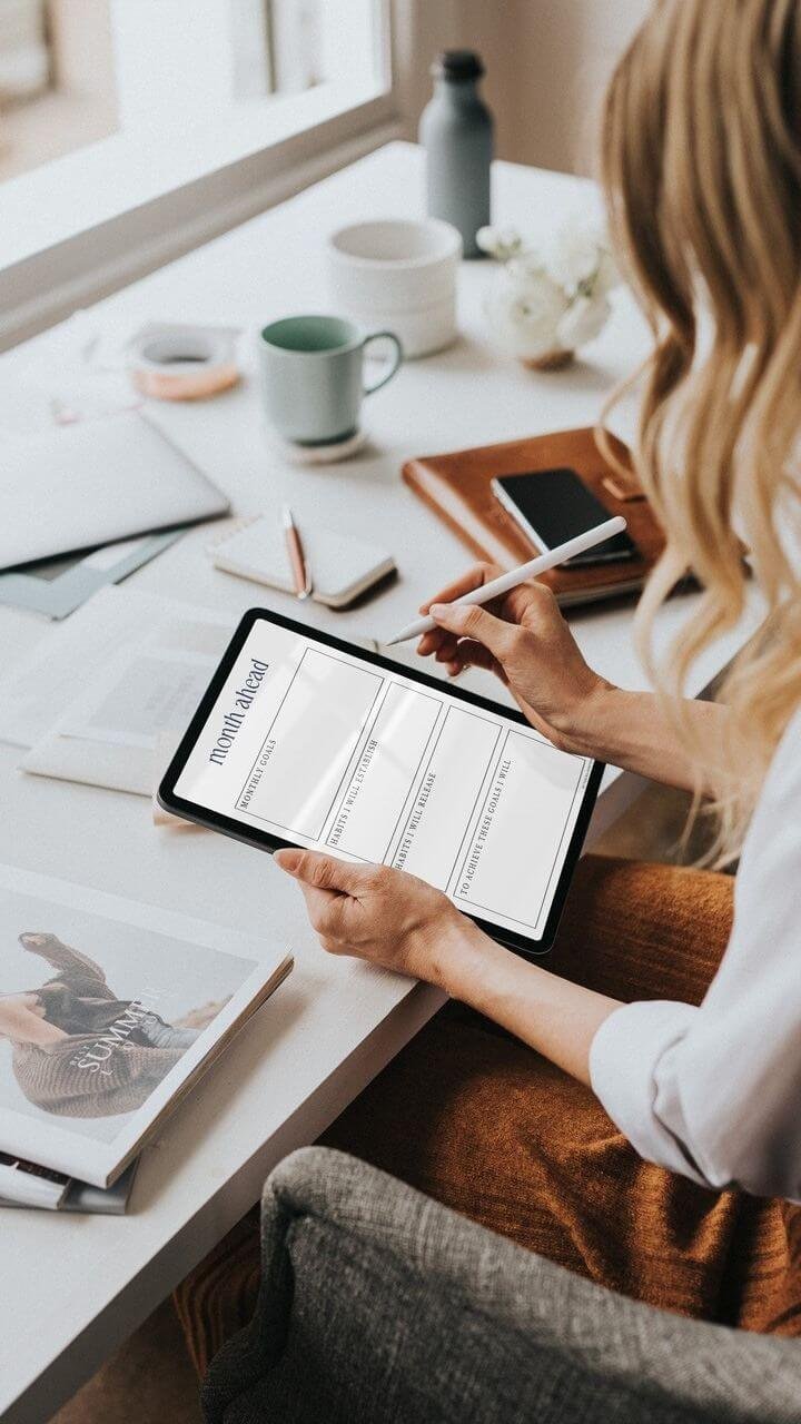 A woman writing in a planner titled 'month ahead' sitting at a desk with a tablet, notebooks, a magazine, a mug, and decorative items in a bright room.