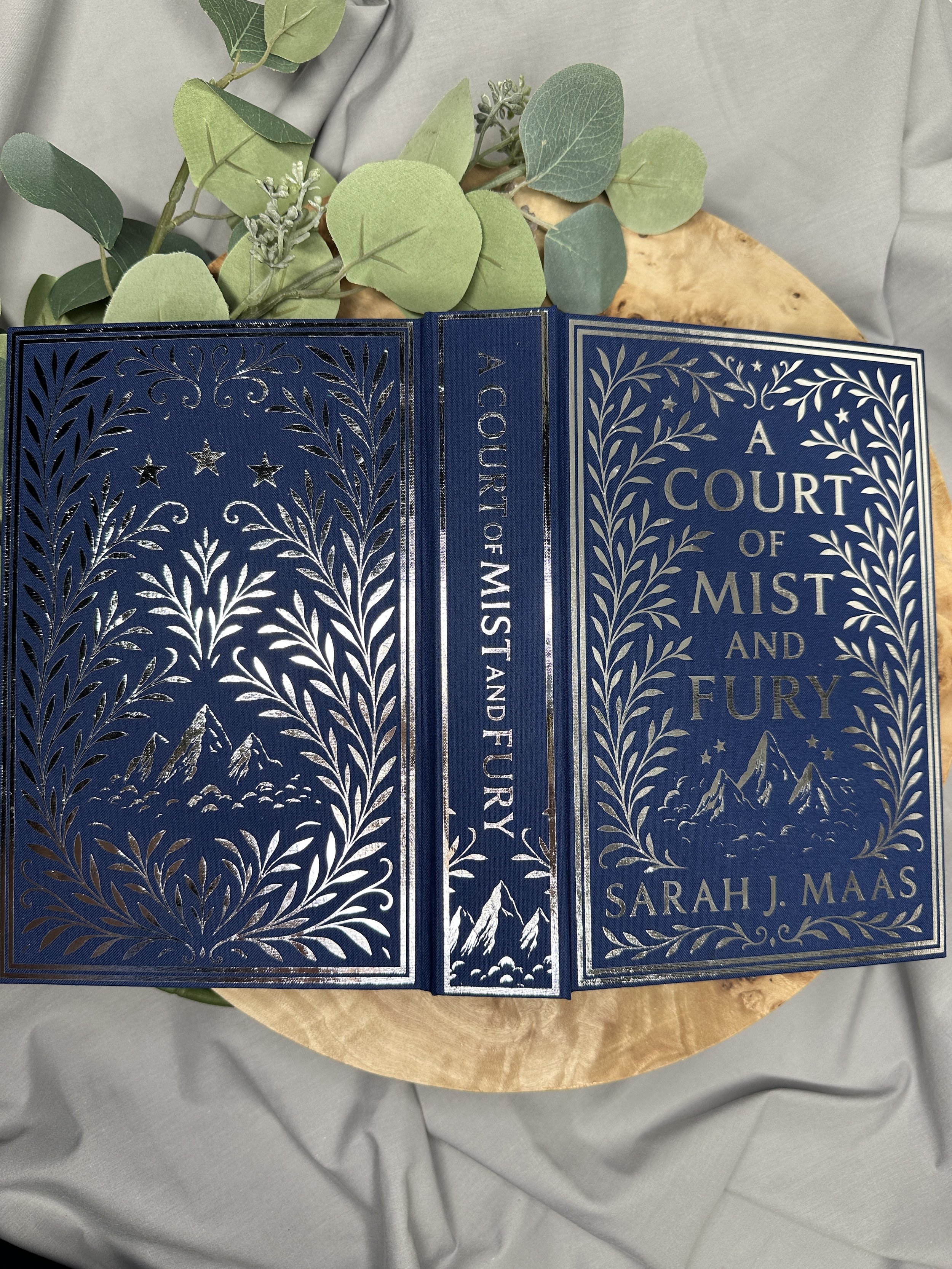 A hardcover book titled 'A Court of Mist and Fury' by Sarah J. Maas, with an intricate silver design of mountain ranges and leafy borders on a blue cover, placed on a wooden surface with eucalyptus leaves nearby.