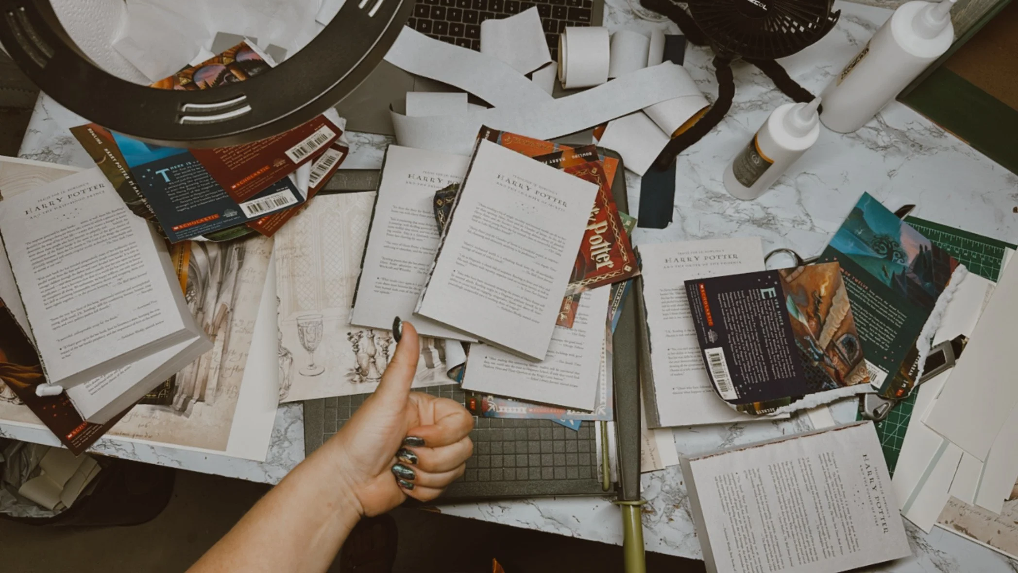 Realistic and chaotic workspace with Harry Potter booklets, printouts, scissors, glue bottles, and torn paper, with a hand giving a thumbs-up.