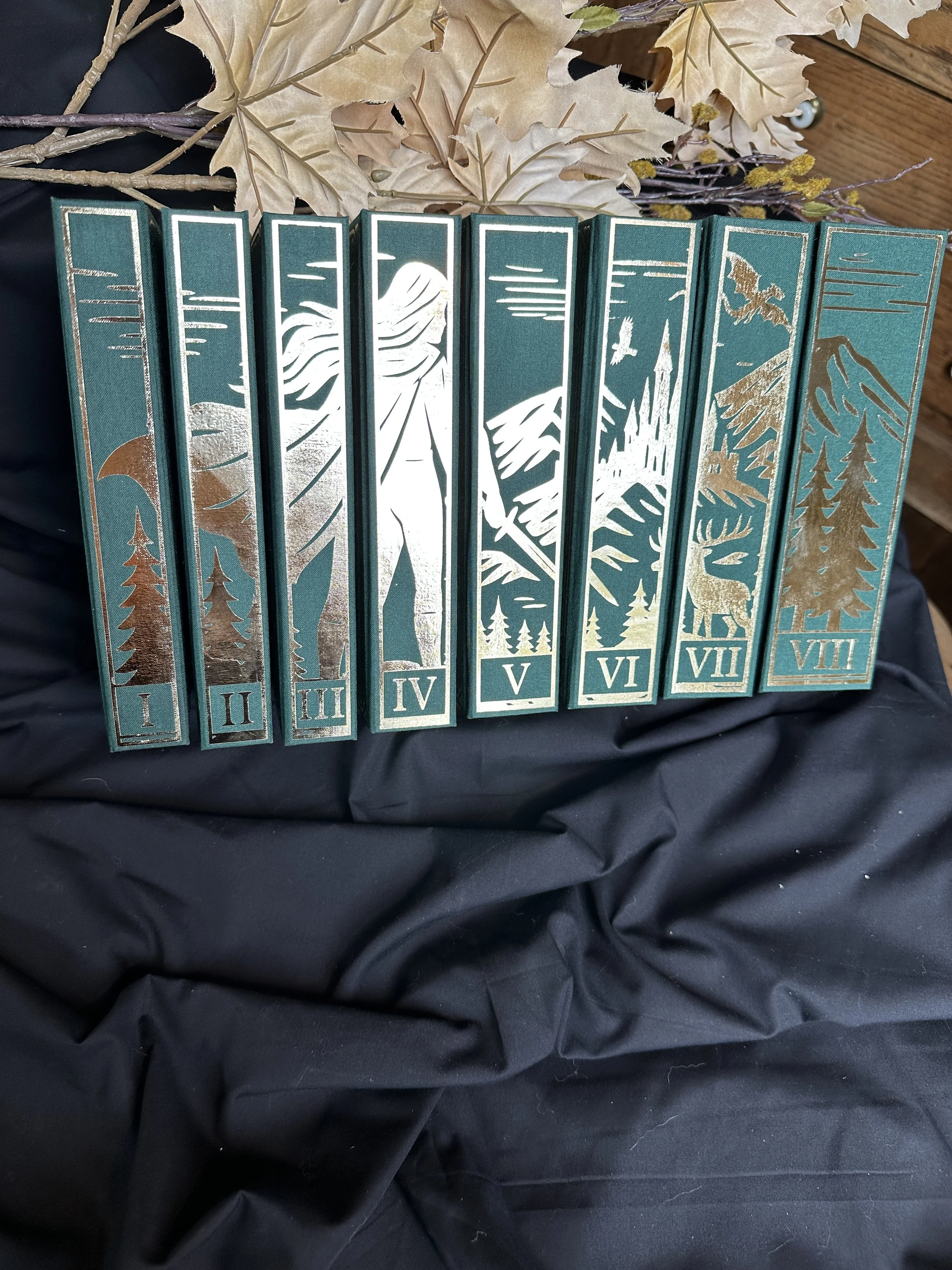 Decorative book spines, when put together depict a whole image with mountain and forest designs, numbered I to VIII, arranged in order on black fabric with autumn leaves and branches in the background.