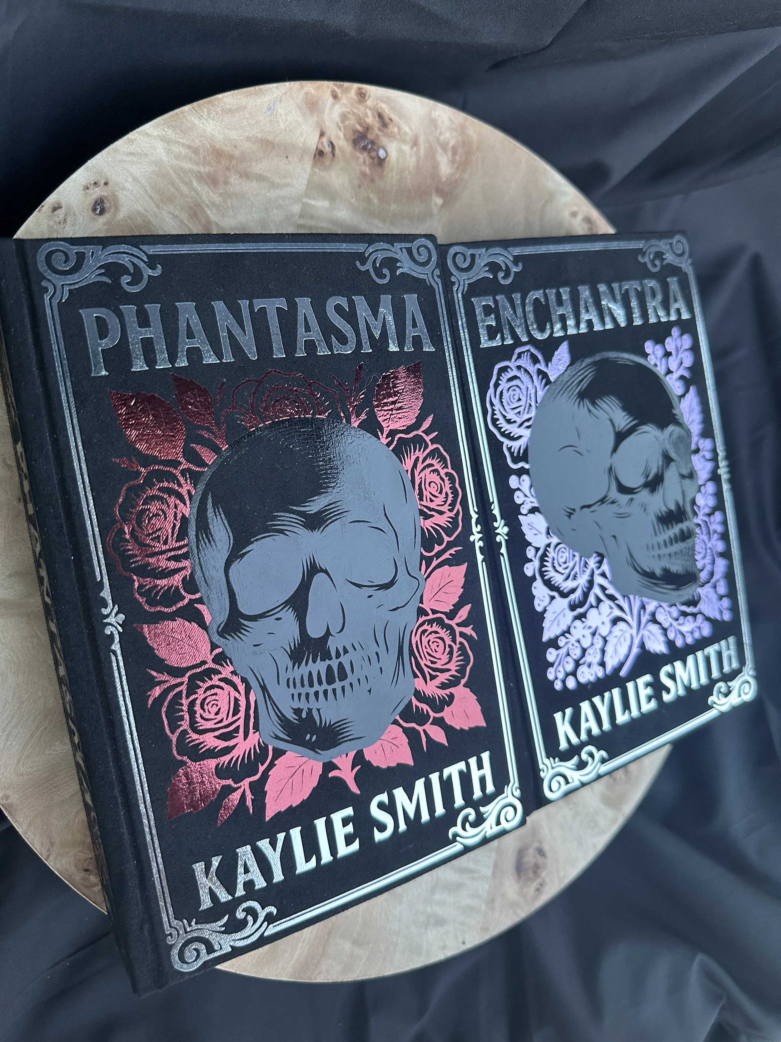 A two book set with titles, "Phantasma" and  "Enchantra" by Kaylie Smith resting on a round wooden table. The book cover features a skull surrounded by roses, with metallic and holographic effects.