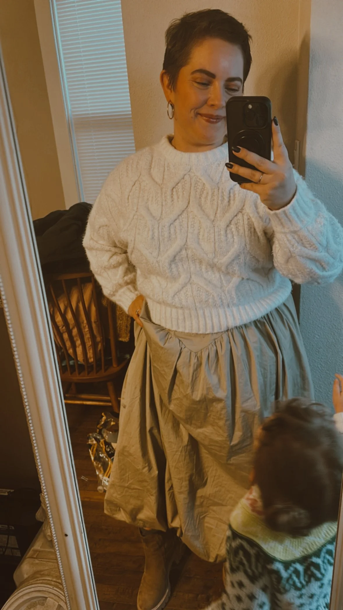 Woman taking a mirror selfie, wearing a white cable-knit sweater, a beige voluminous skirt, brown boots, and earrings, with a child partially visible in the foreground.