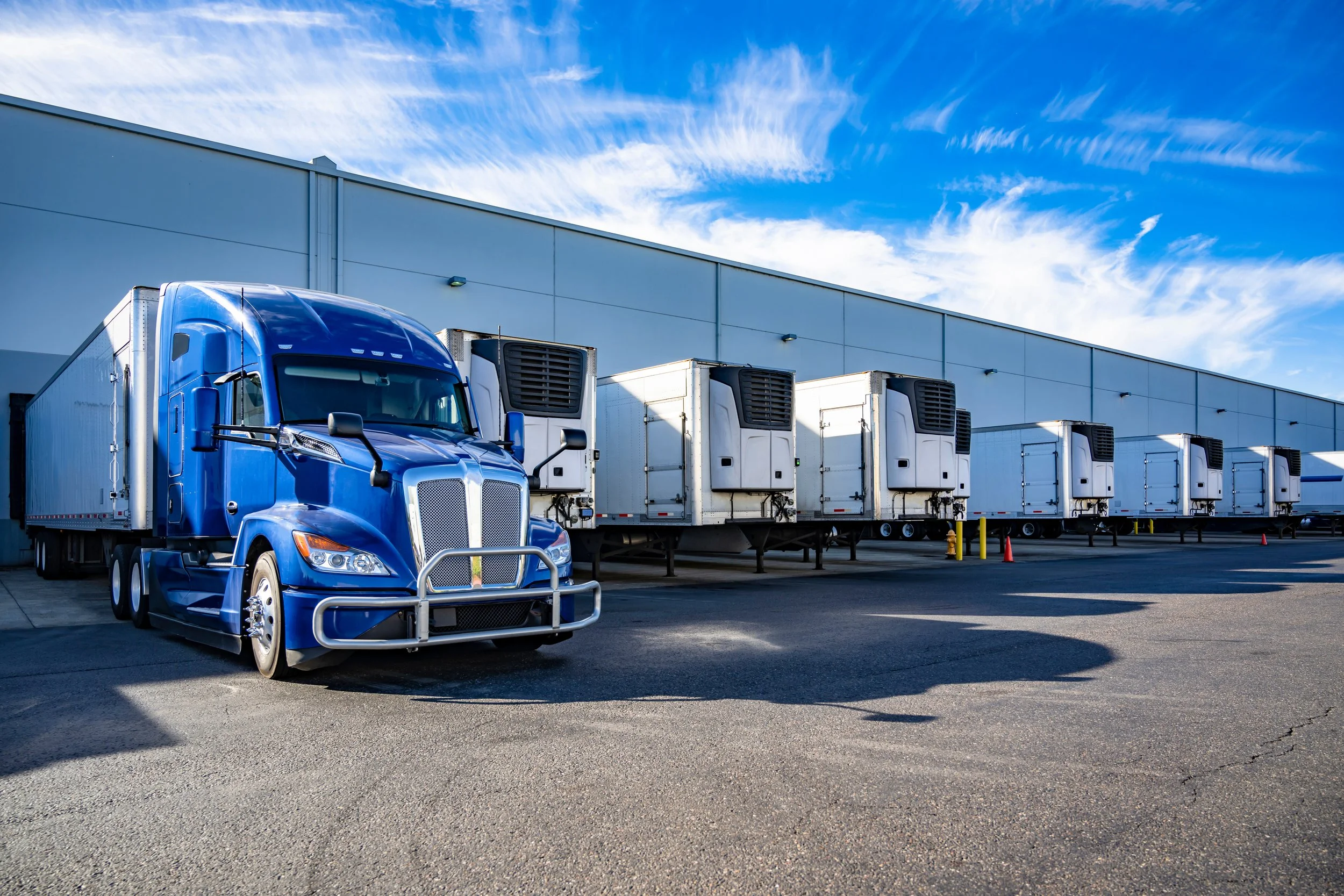 Blue semi-truck with multiple refrigerated trailer units parked outside a large warehouse with a clear sky.