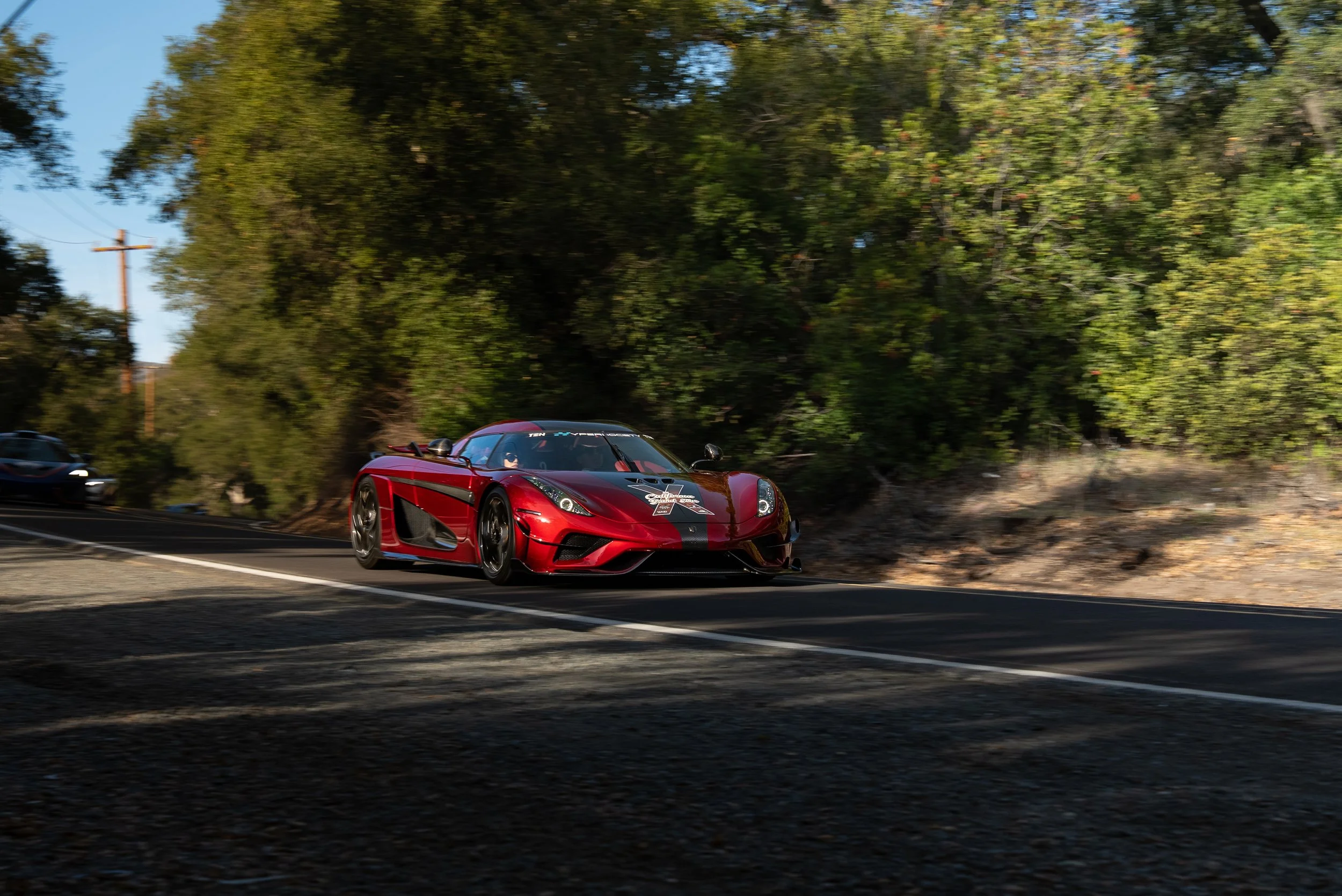 Red sports car with black racing stripes driving on a winding mountain road surrounded by green trees.