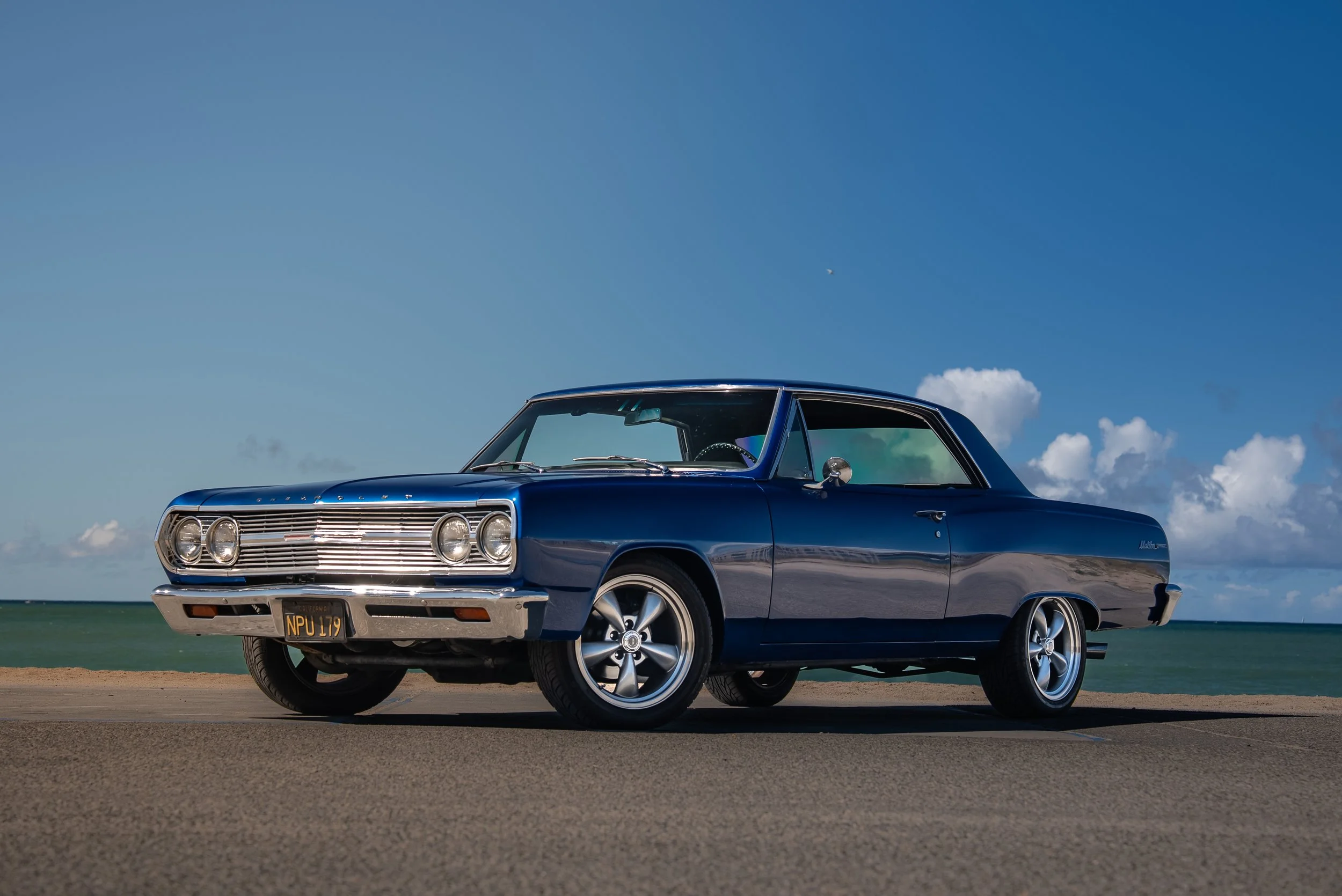 A vintage blue two-door car parked on a beachside road with the ocean and partly cloudy sky in the background.