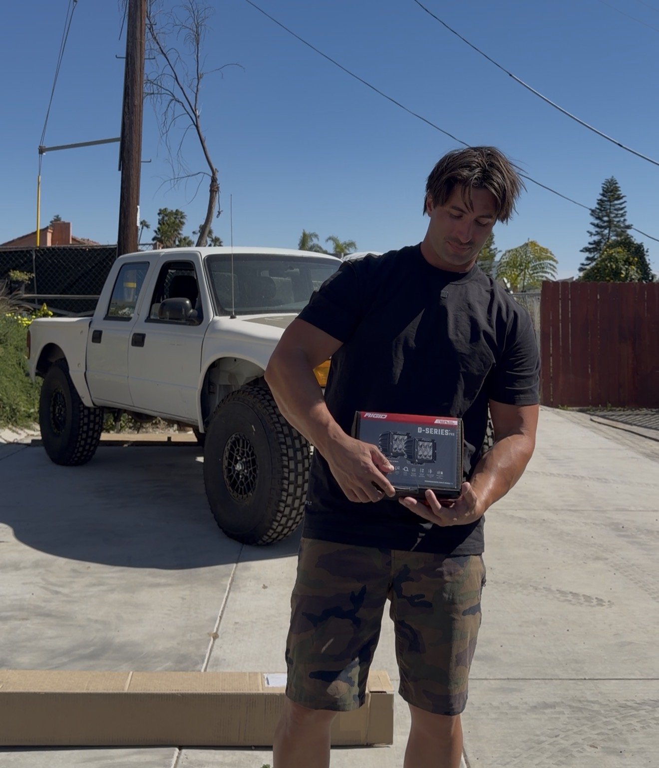 A young man with brown hair, wearing a black T-shirt and camouflage shorts, stands outdoors on a concrete driveway holding a box of computer components. Behind him is a white pickup truck parked near a wooden fence, with trees and a clear blue sky in the background.