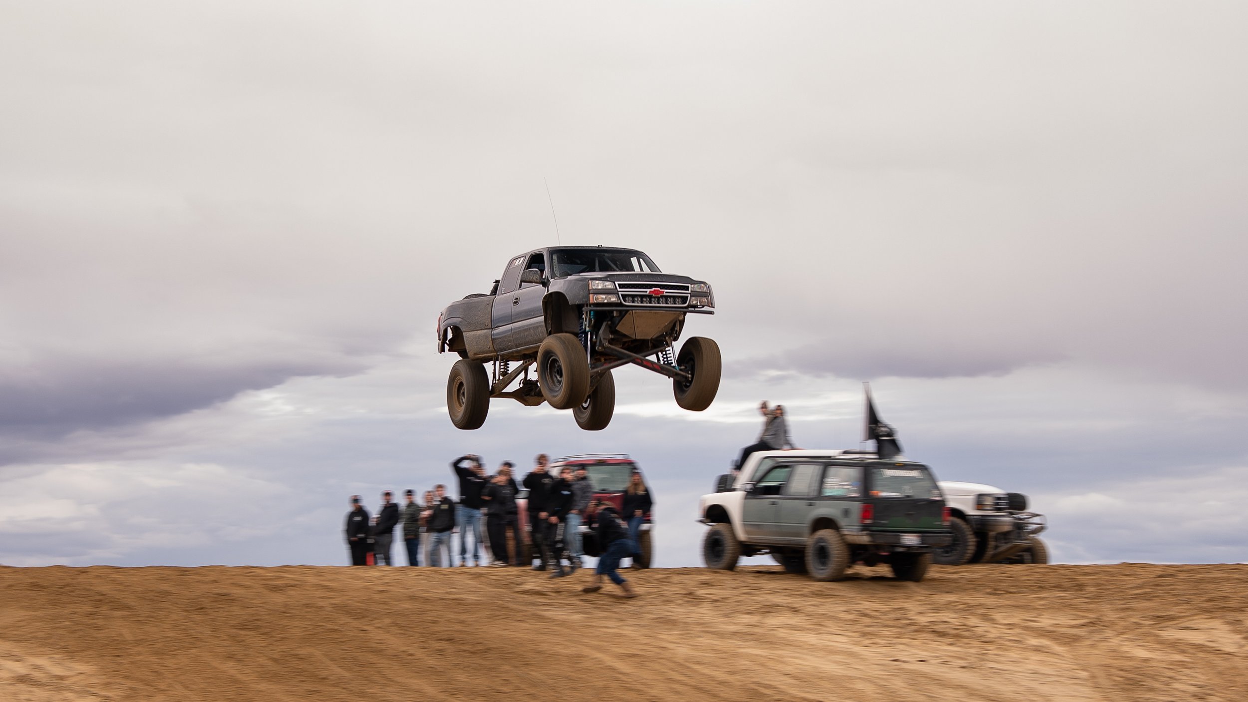 A modified pickup truck is airborne above a dirt track during a rugged vehicle stunt, with a group of spectators and support vehicles in the background under a cloudy sky.